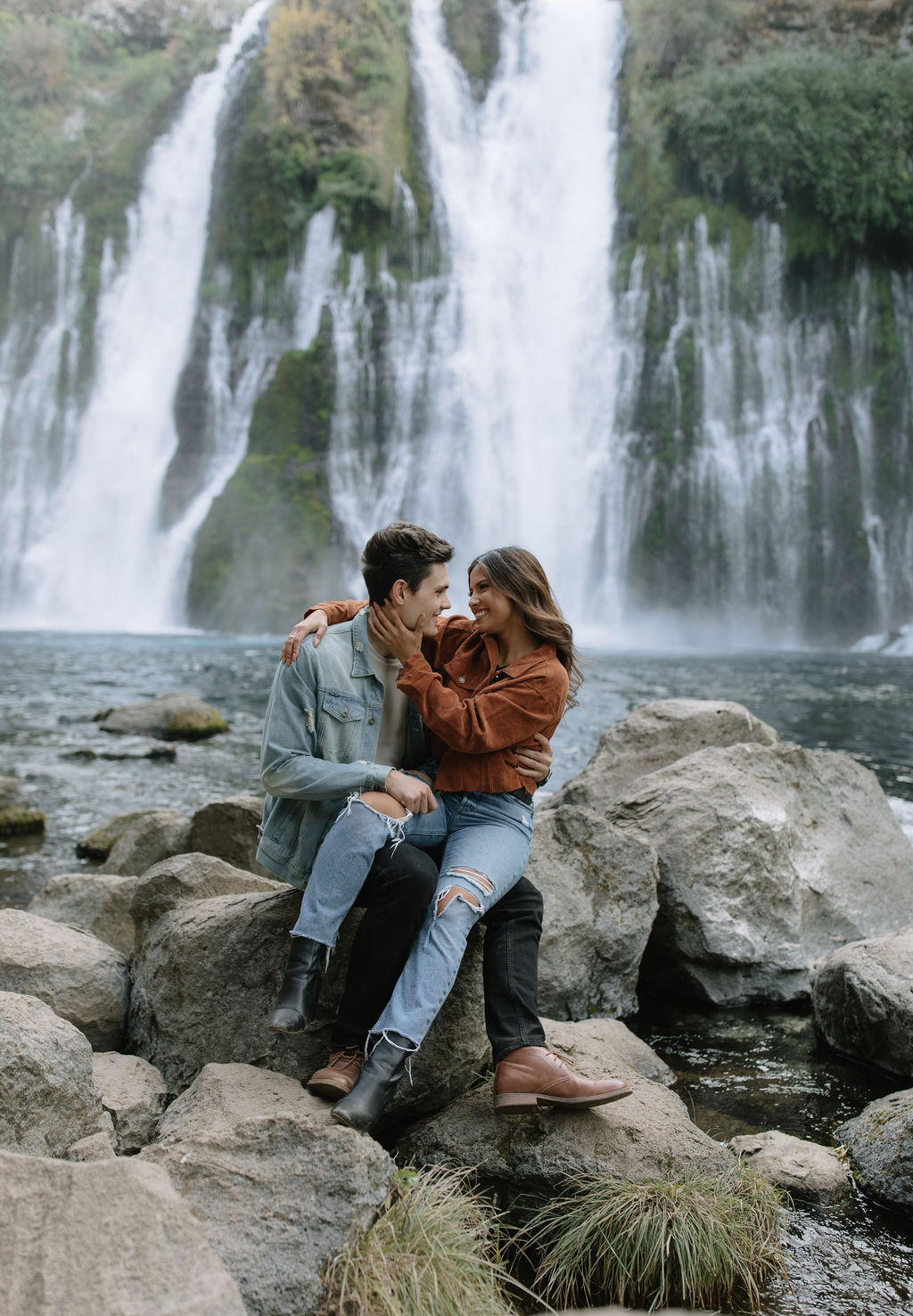 Couple smiling at each other sitting on the rocks in front of waterfalls