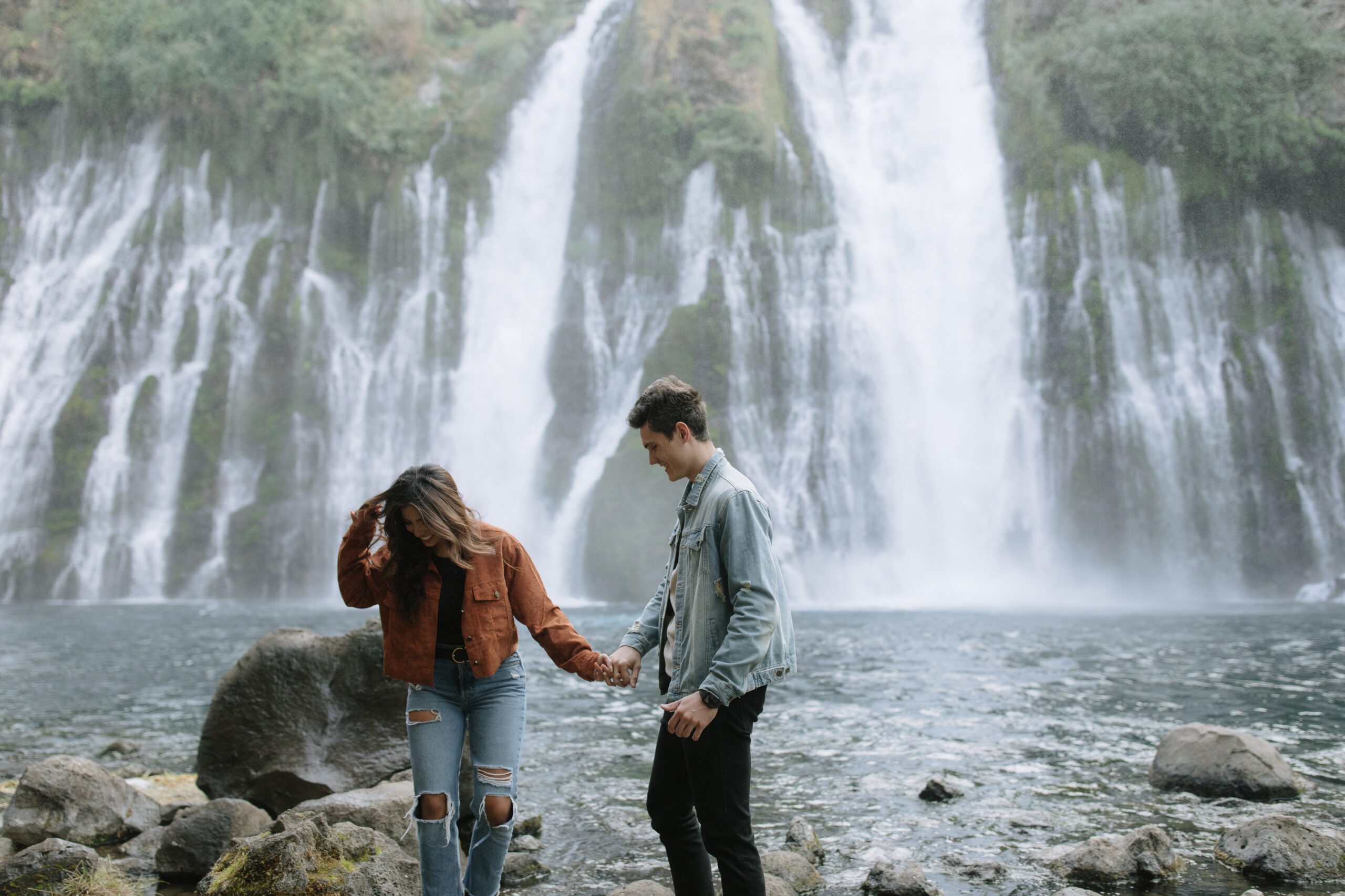 Couple holding hands walking in front of burney falls waterfalls