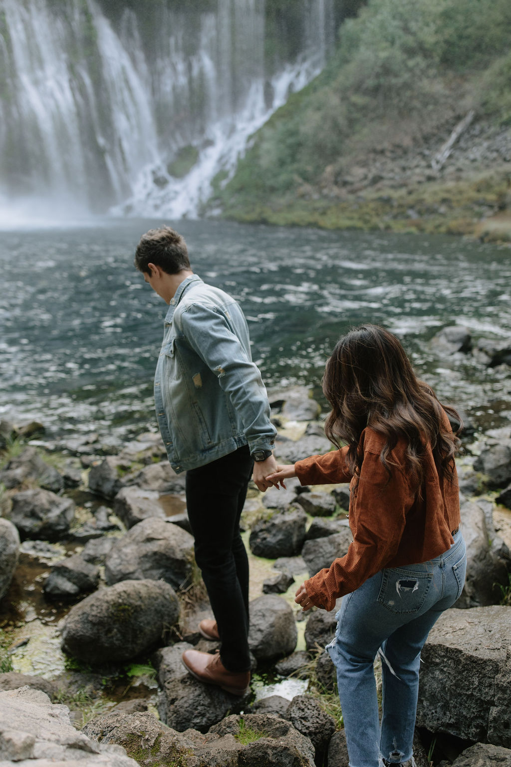 Couple holding hands walking towards waterfalls at Burney Falls