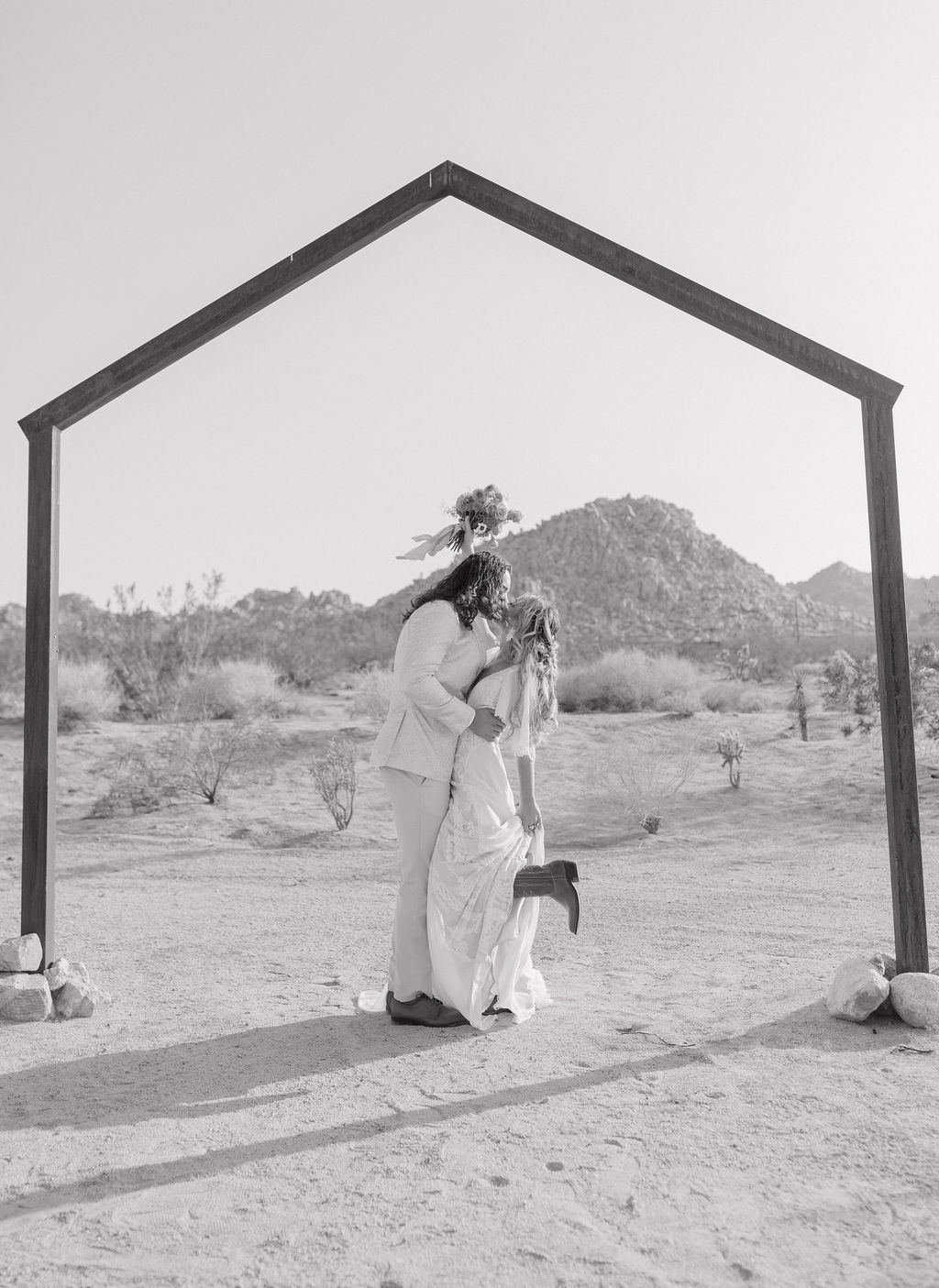 A black and white photo of a bride and groom kissing underneath a wooden wedding arch