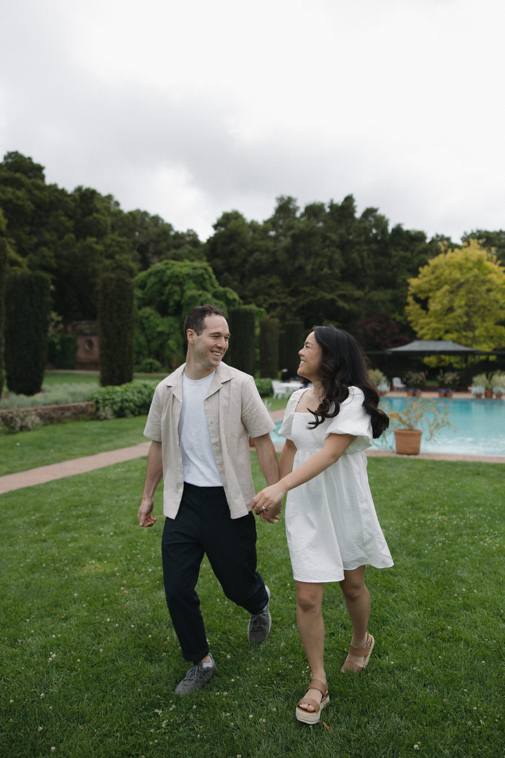Couple walking holding hands and smiling at each other in front of pool at engagement photoshoot