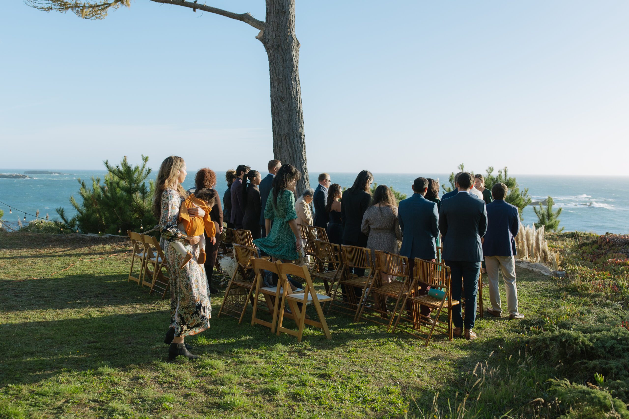 Intimate wedding ceremony photo of guests standing while bride walks down the aisle with the ocean in the background