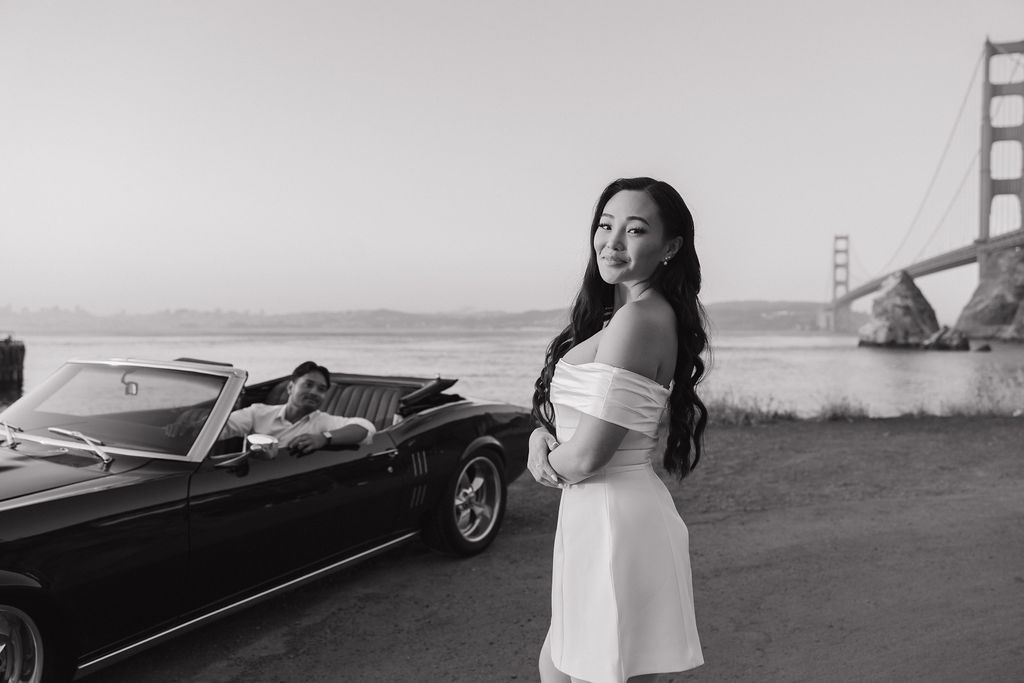 Bride standing next to car while groom looks at her from it in black and white engagement photo