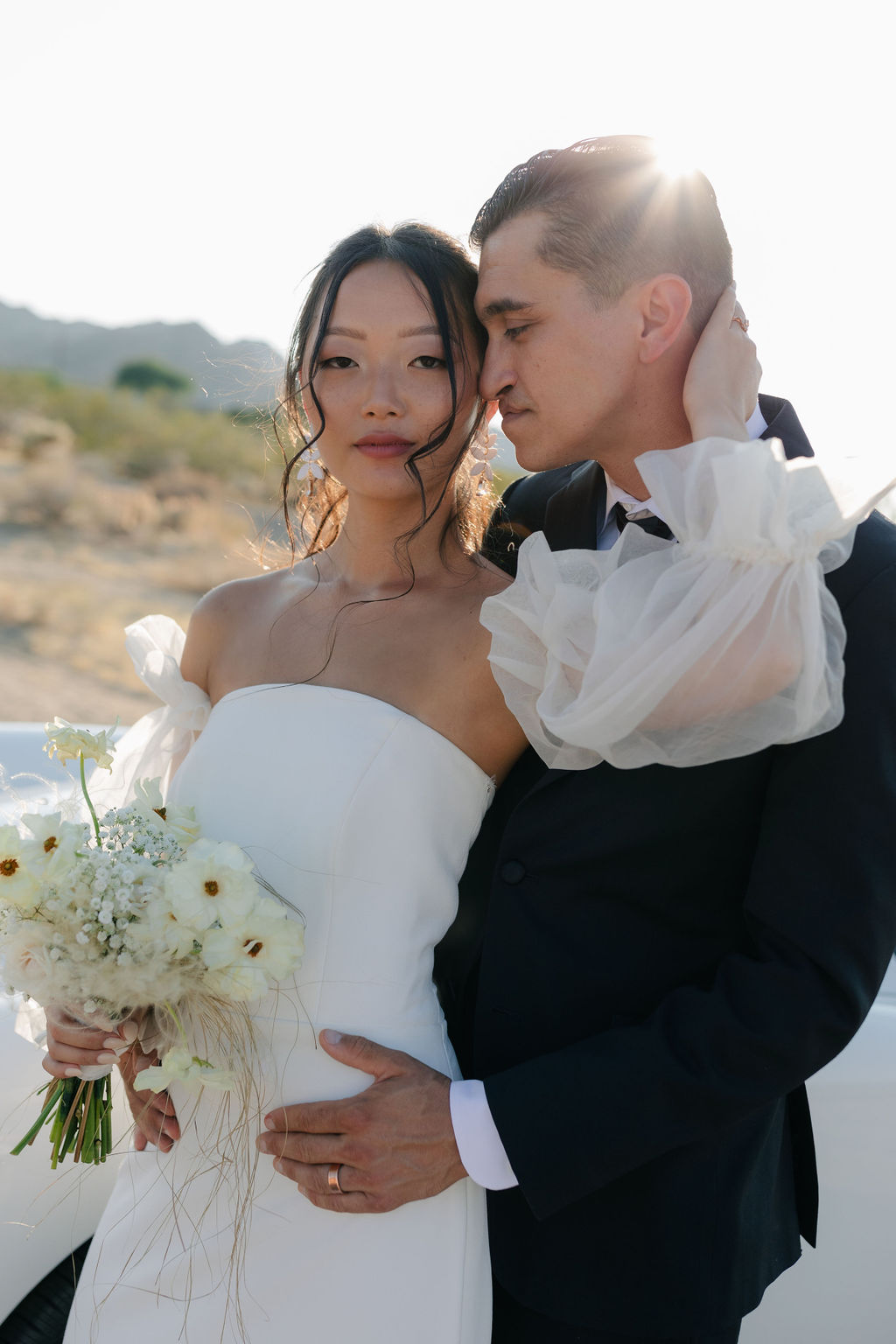 Bride holding groom's head with sun hitting in the back and a vintage car from Joshua Tree wedding