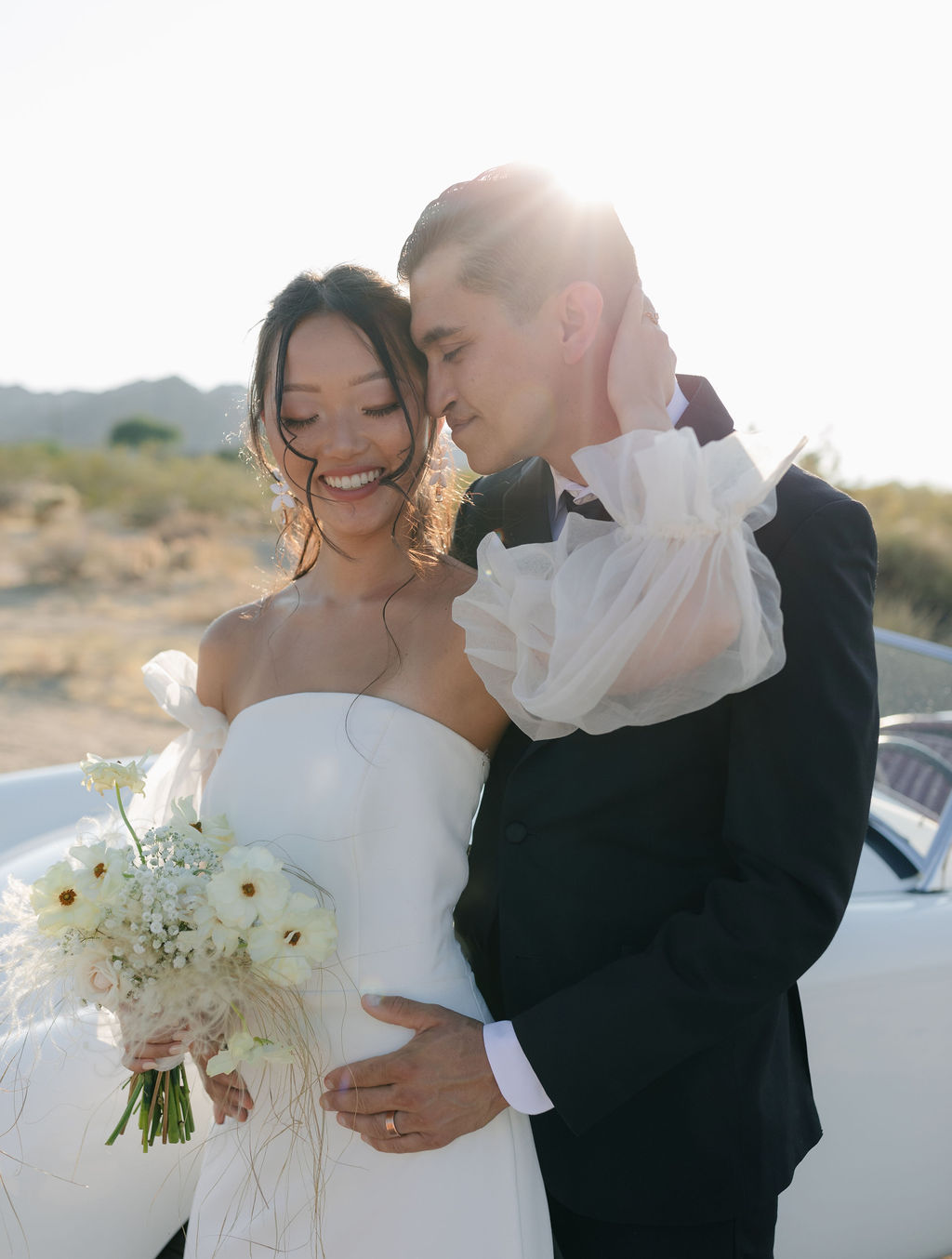 Sunny Joshua tree wedding photo with white wedding bouquet and vintage car with bride's hand on groom's face