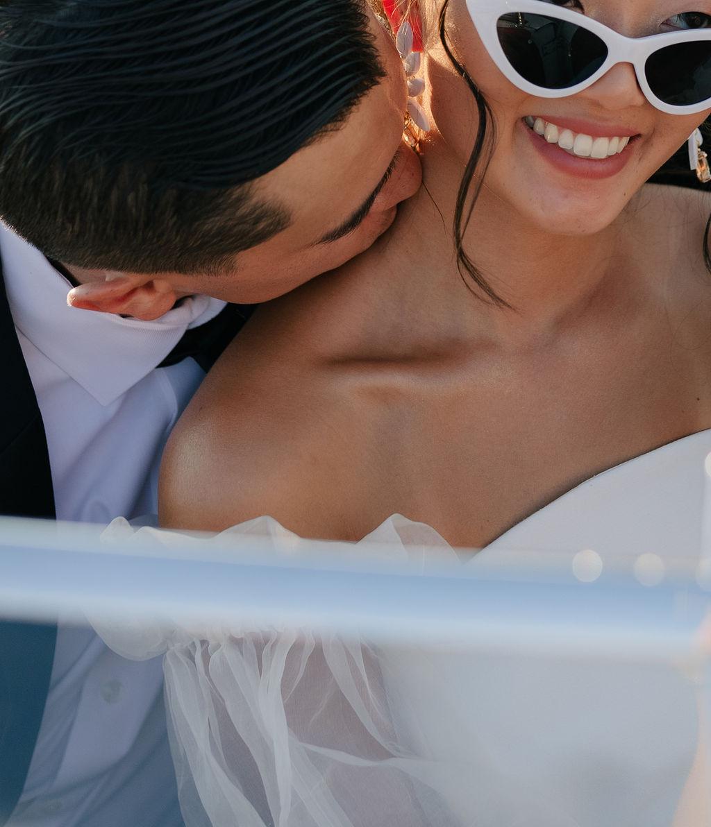 Up close photo of bride and groom with bride wearing sunglasses and groom kissing her on the neck