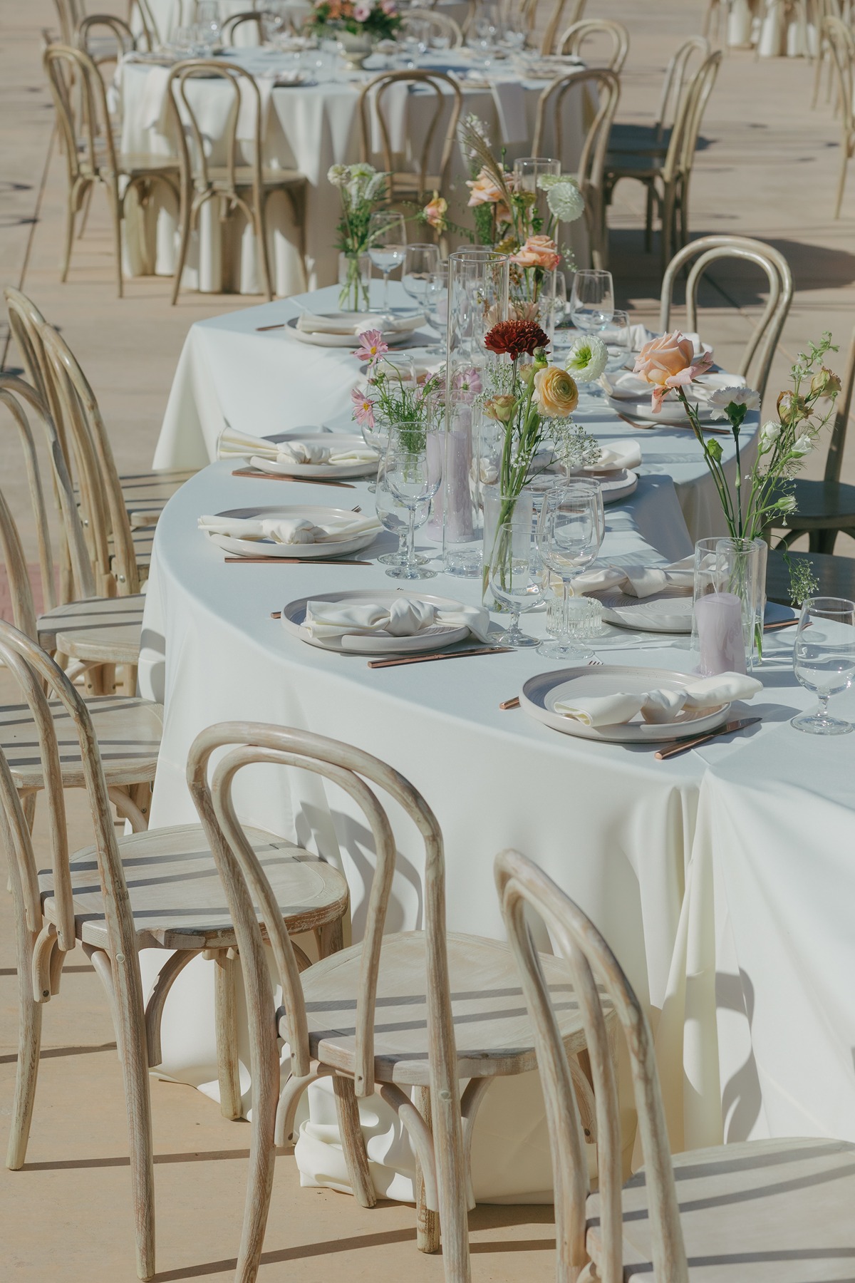 Snaked table setup for wedding reception at luxury wedding with pastel and red wildflowers and ivory tablecloths with wooden chairs