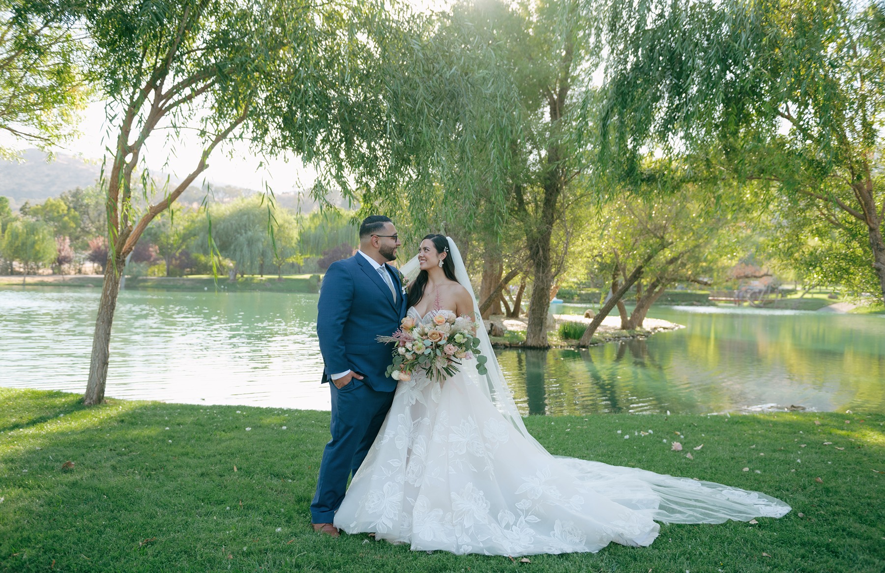 Bride and groom smiling at each other in front of lake during wedding photos