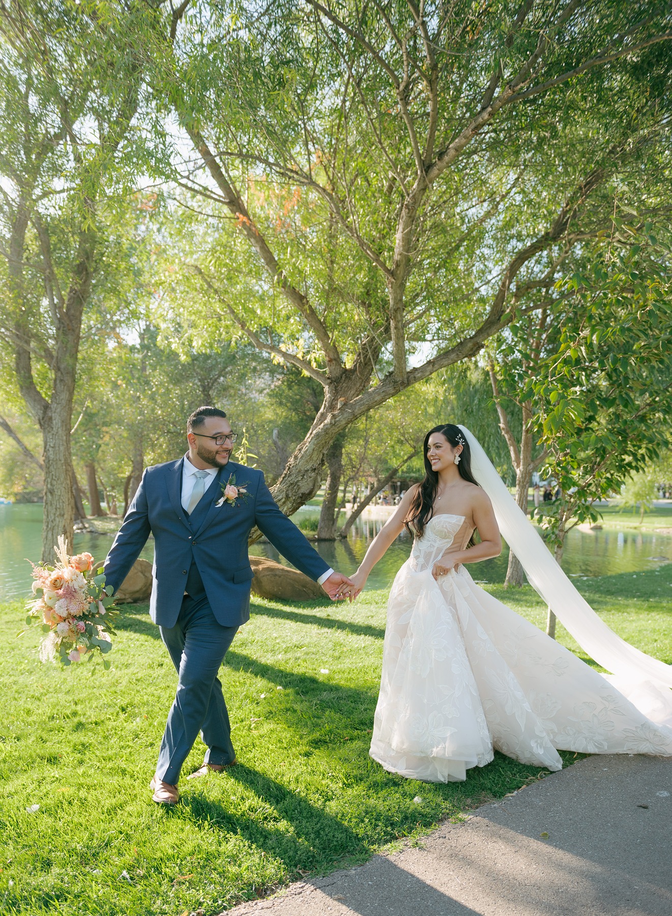 Bride and groom holding hands walking through a field with the sun and a big tree behind them