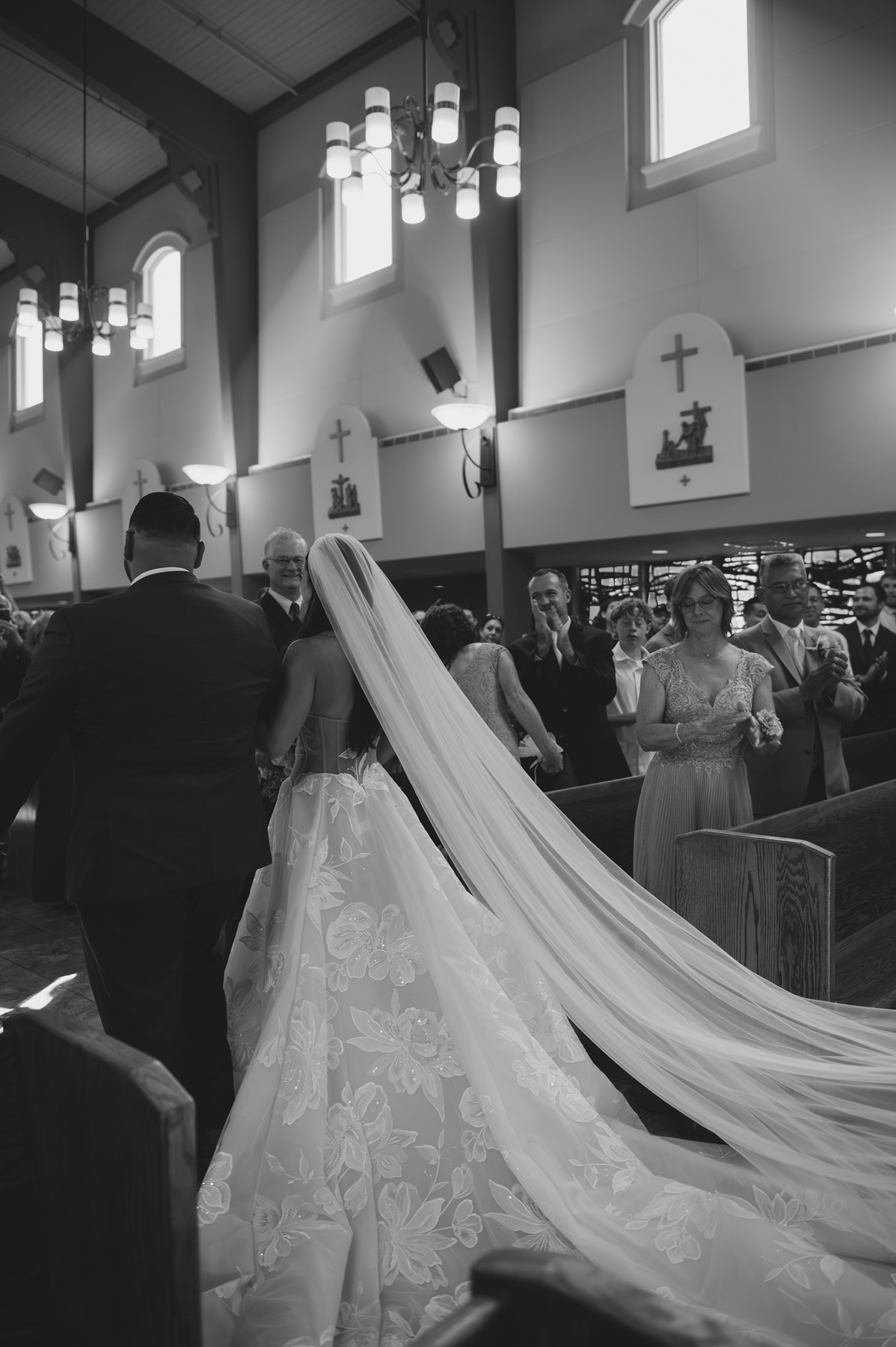 Black and white photo of bride and groom walking down the wedding aisle with cathedral veil trailing behind bride
