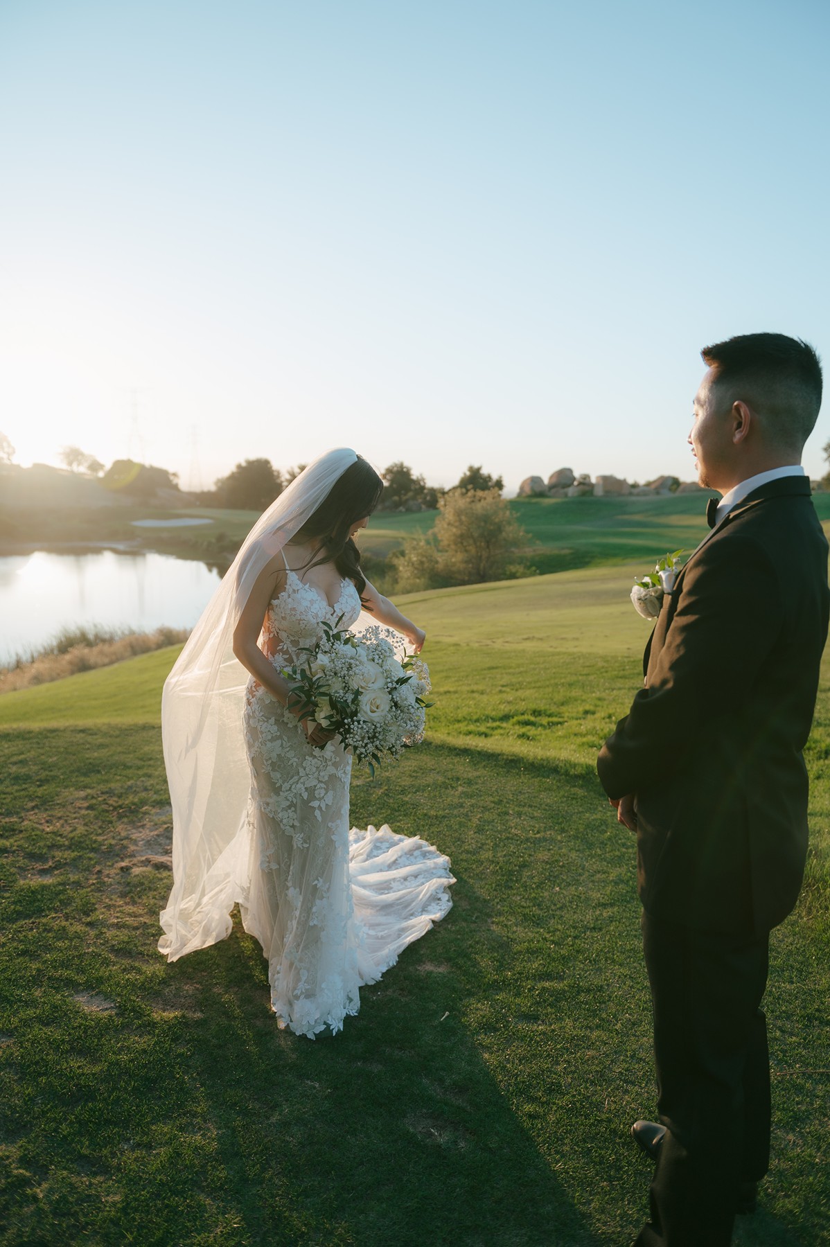 Bride holding her white bouquet and looking down at her veil while groom looks at her