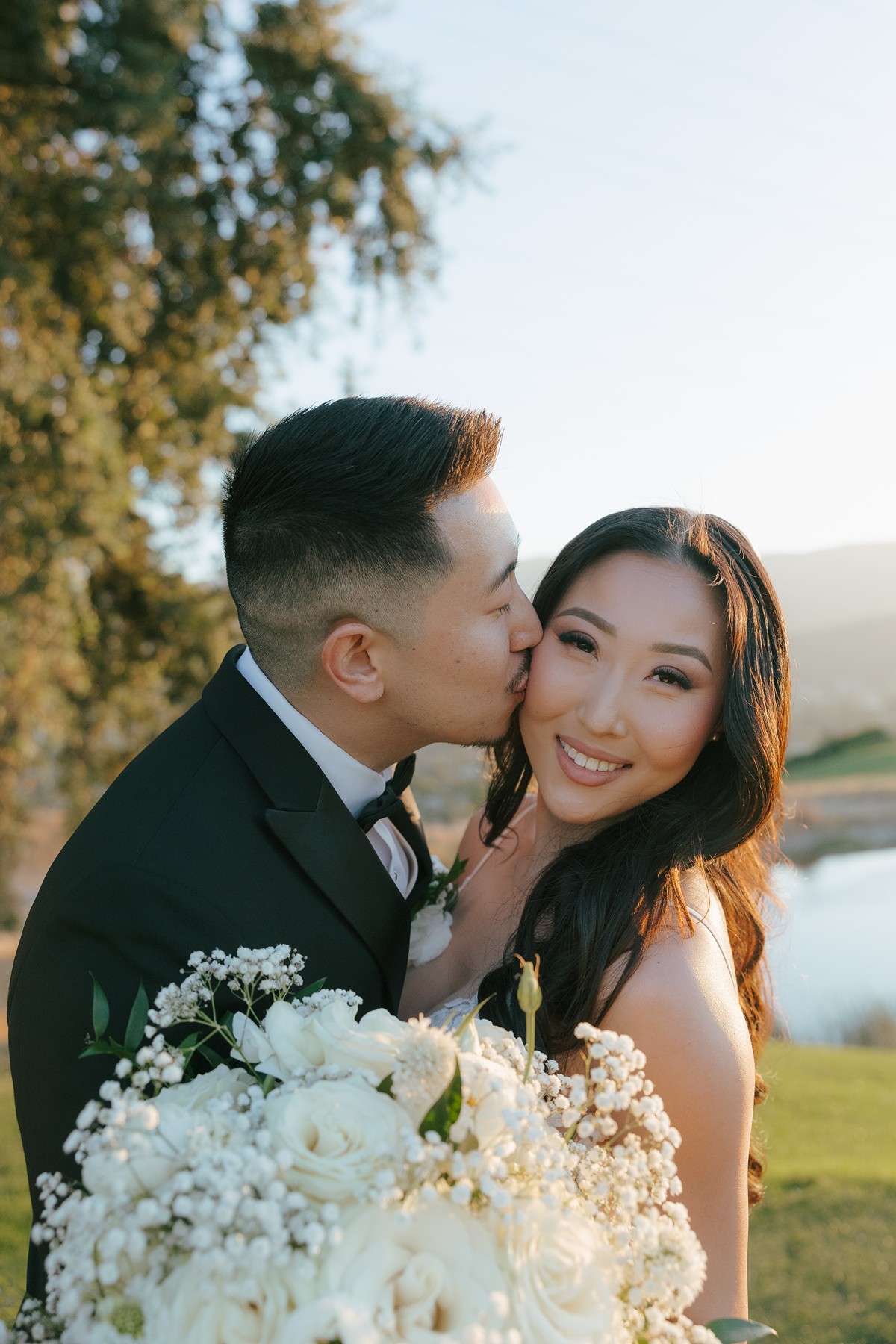 Bride and groom posing for couples photos with the bride smiling at the camera while the groom kisses her cheek