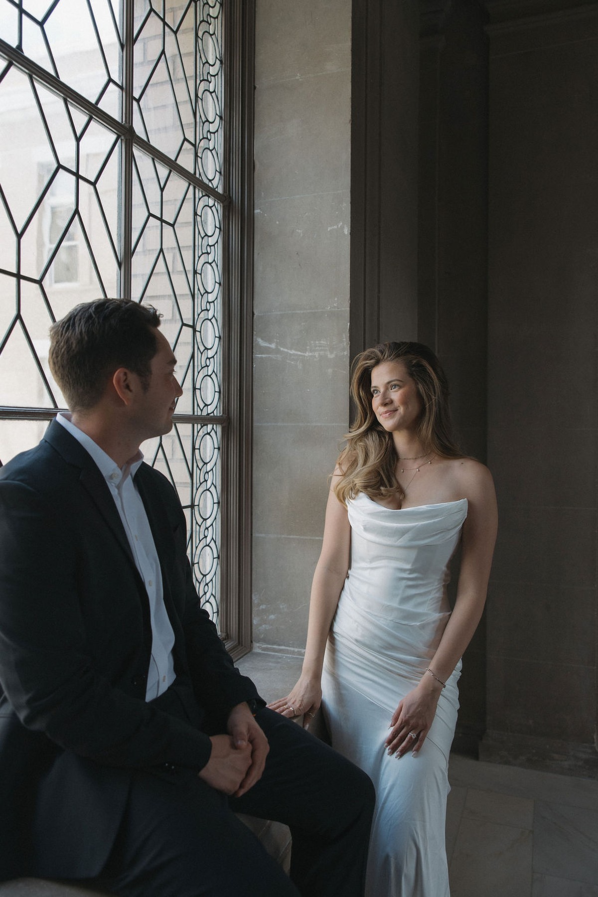 Couple posing on window sill with the bride smiling at the groom who is sitting down on the windowsill