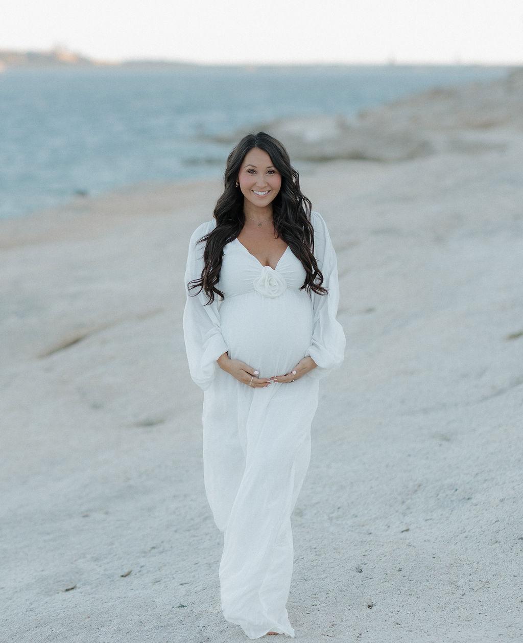 A woman wearing a white dress with a flower and holding her baby bump, smiling for a photo