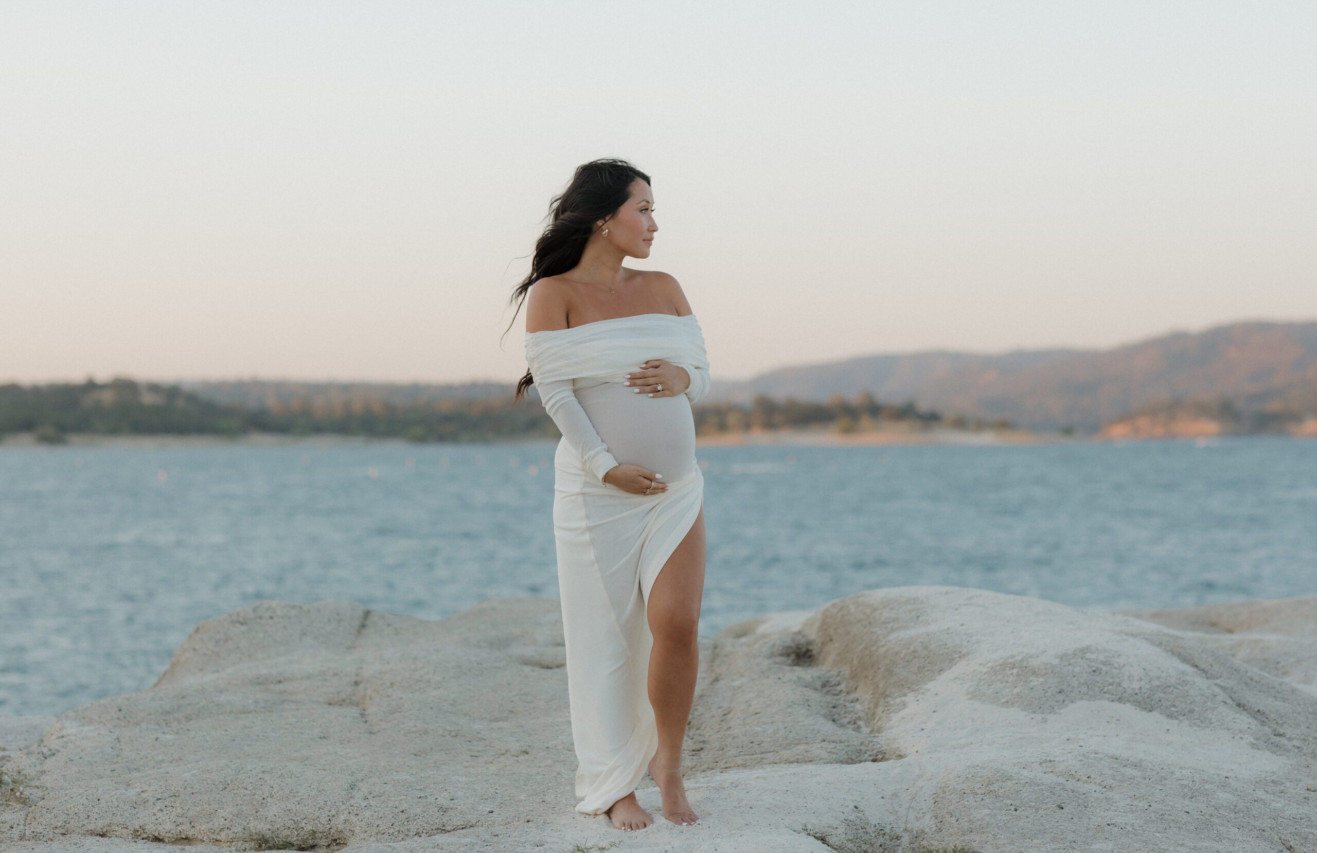 A woman holding her baby bump in a tight-fitted white dress in front of a lake for her maternity photoshoot