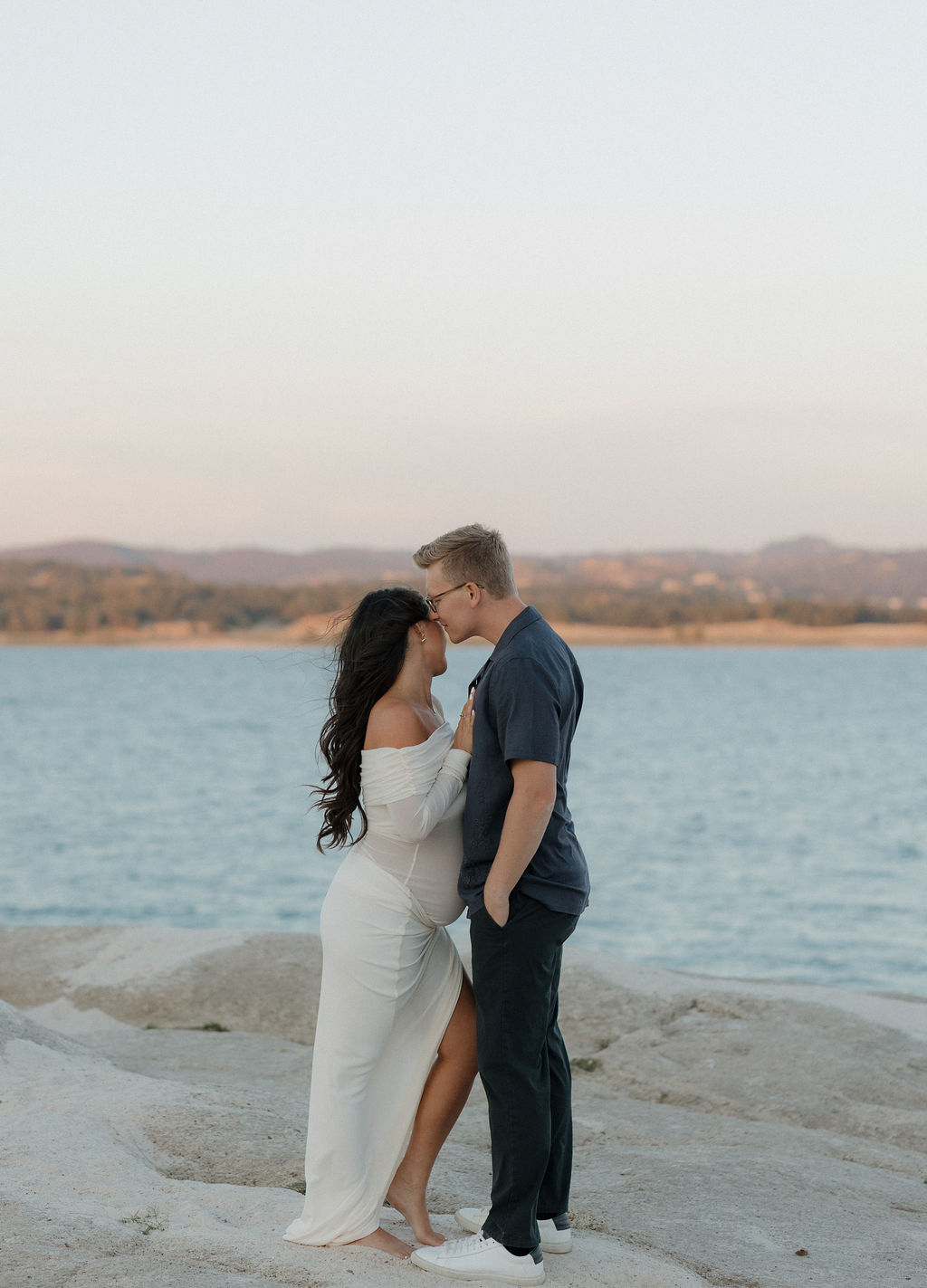 A couple standing to pose in front of Folsom Lake for their maternity photoshoot