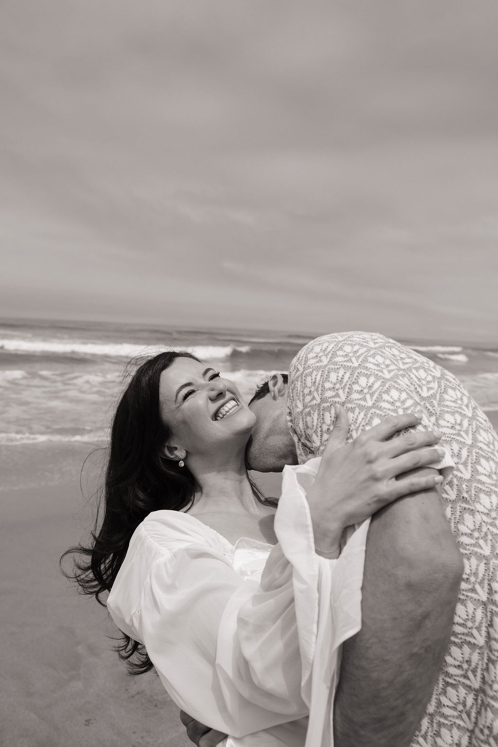 Black and white beach engagement photo of man kissing his fiancee on the neck