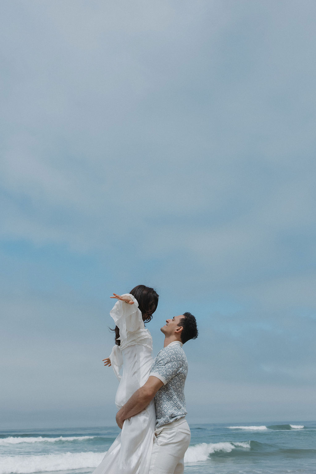 Man holding his fiancee up and her putting her arms out on the beach