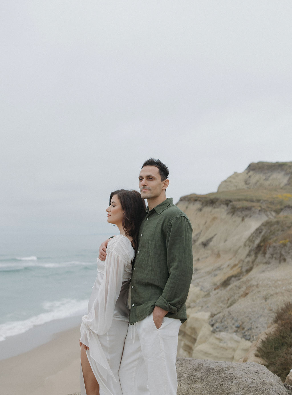 Woman leaning on man on a cliffside during beach engagement photos