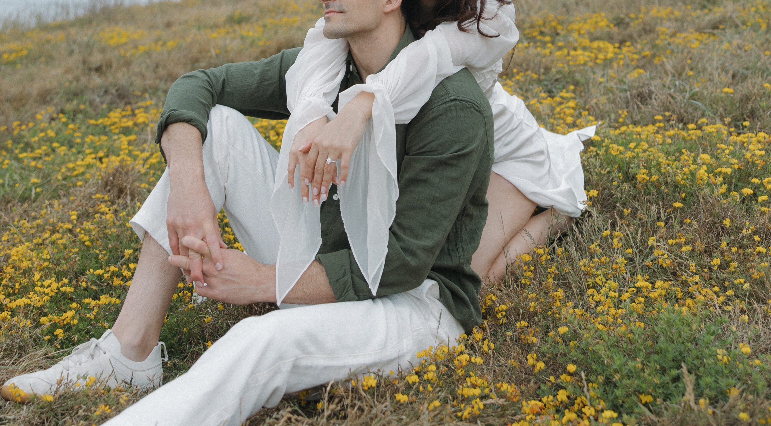 Woman hugging her fiancé from behind and showing off her engagement ring in a flower field