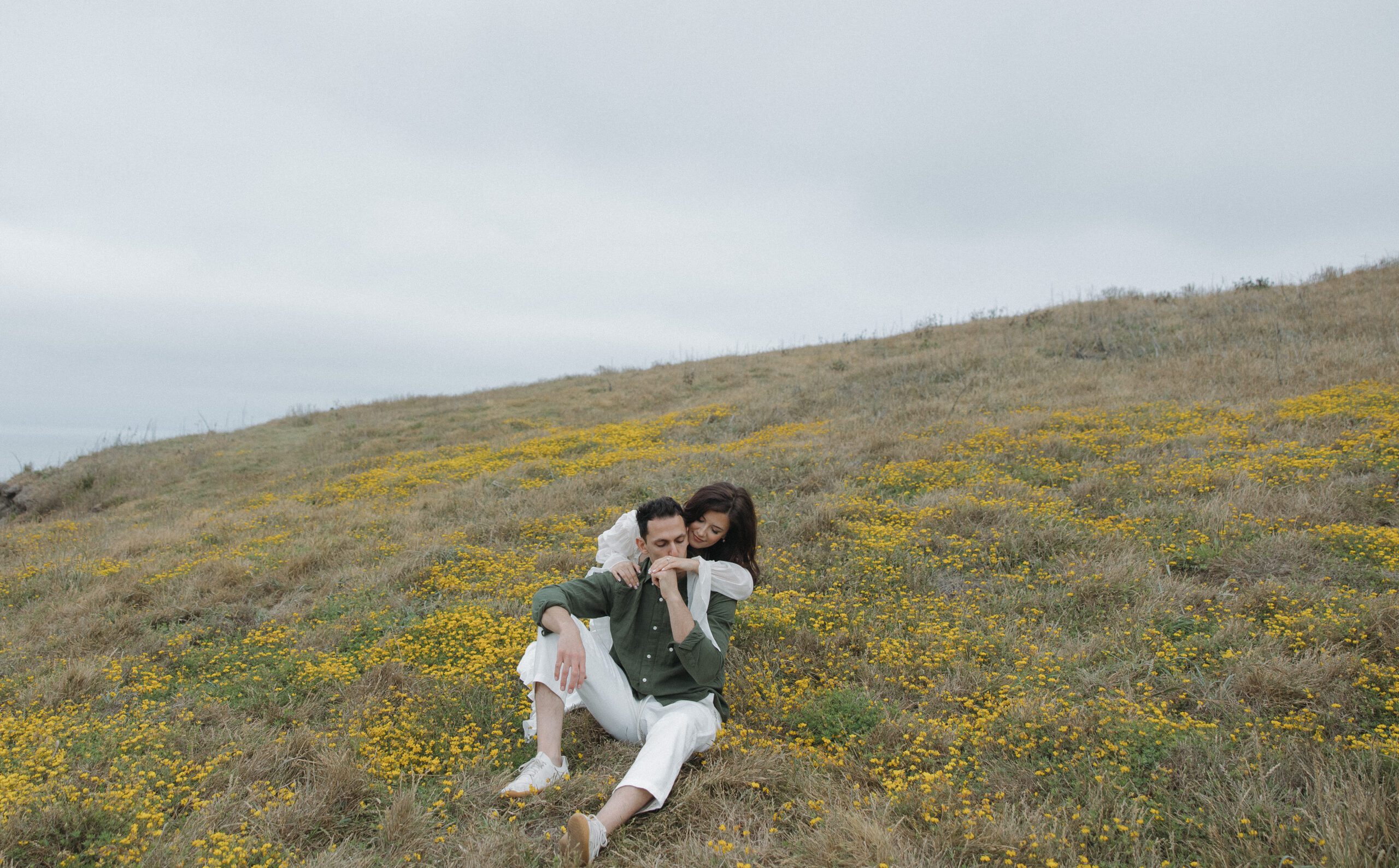 Couple sitting in a field with yellow flowers, with man kissing woman's hand