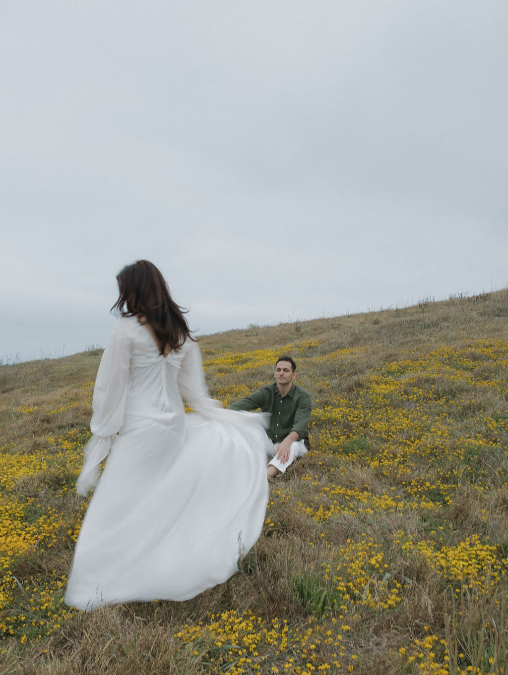 Woman spinning in a white flow dress while her fiancé sits in a field watching her