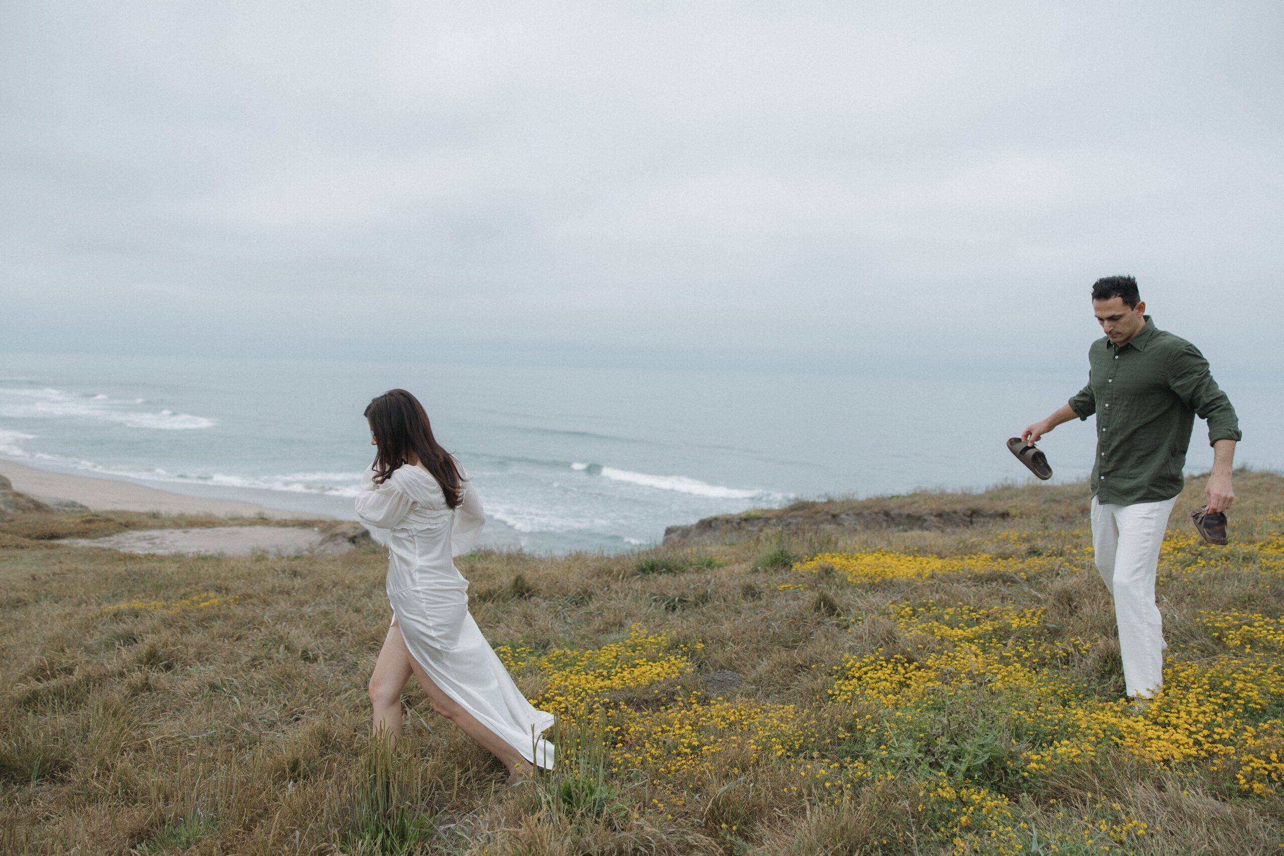 Candid engagement photo of couple walking through a field on a cliff in California