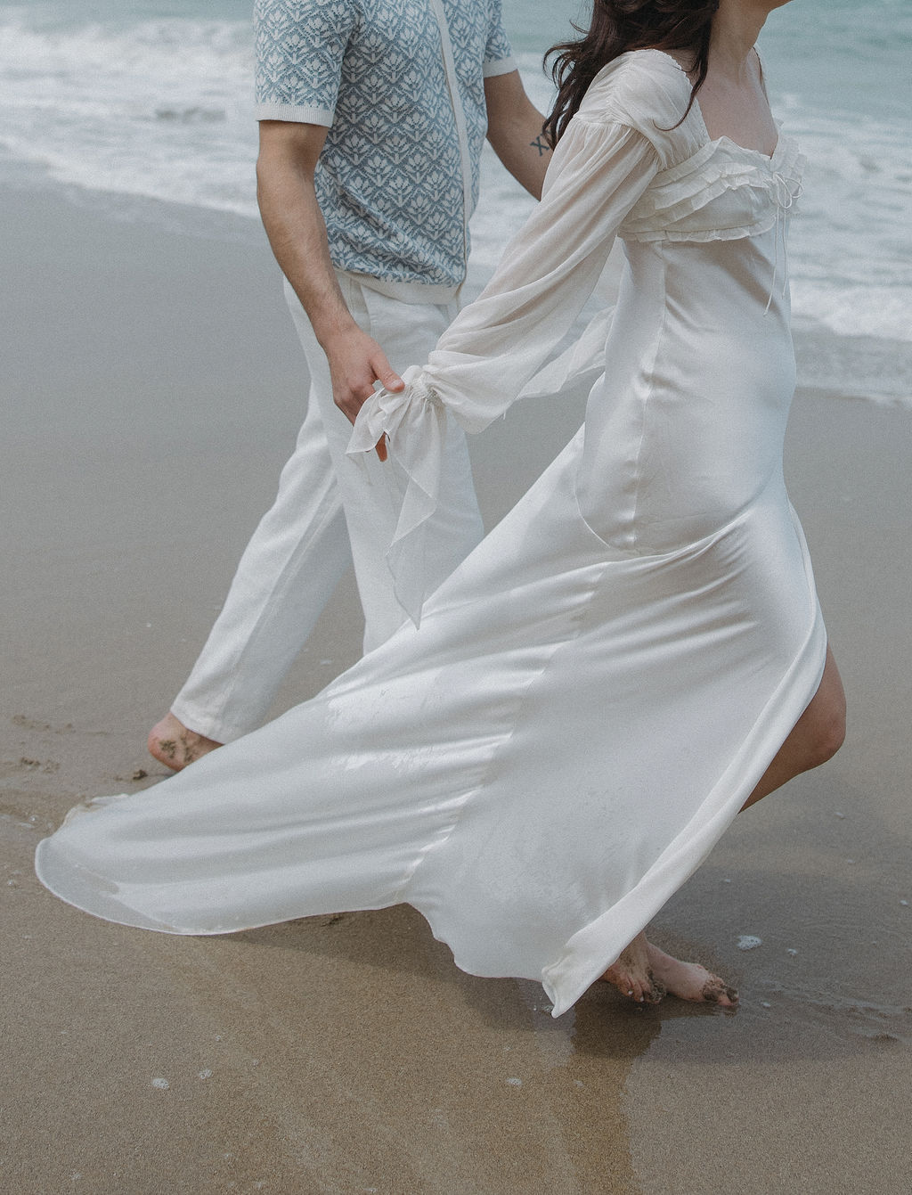Editorial engagement photo of couple walking on the beach either man holding woman's hands walking behind her