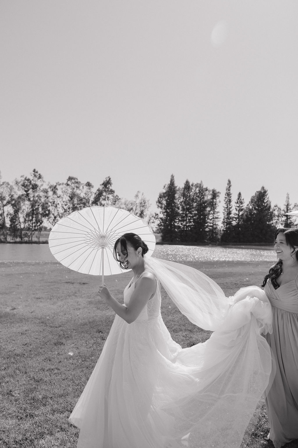 Black and white photo of maid of honor carrying bride's dress while bride holds parasol