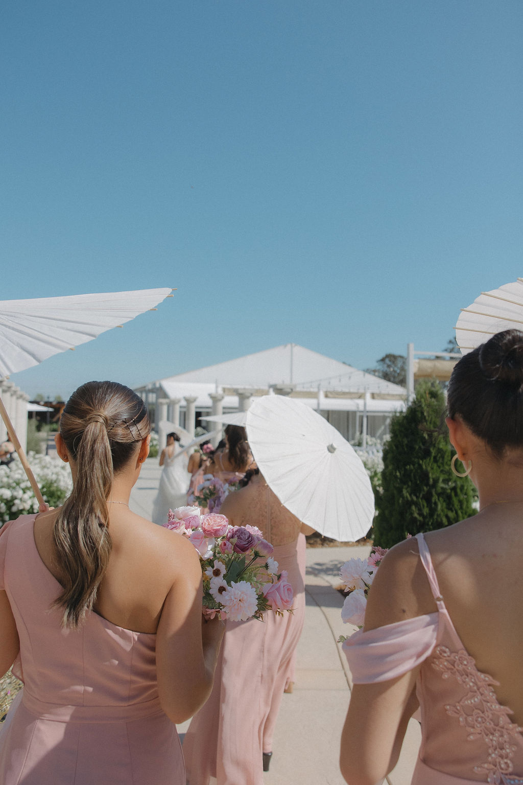 Bridesmaids in pink bridesmaid dresses carrying parasols while walking behind the bride
