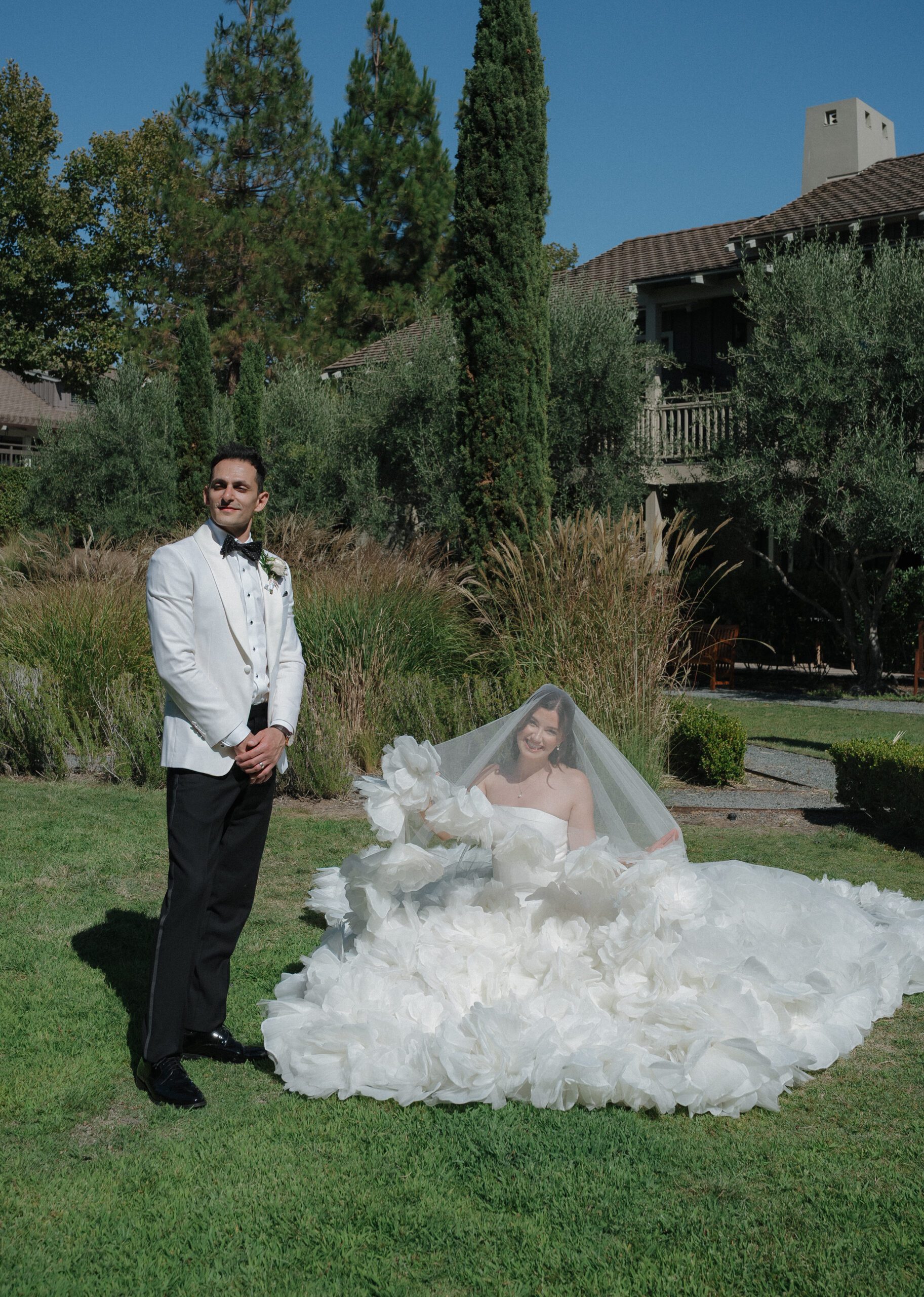 Bride sitting on the ground on a lawn while groom looks away from the camera