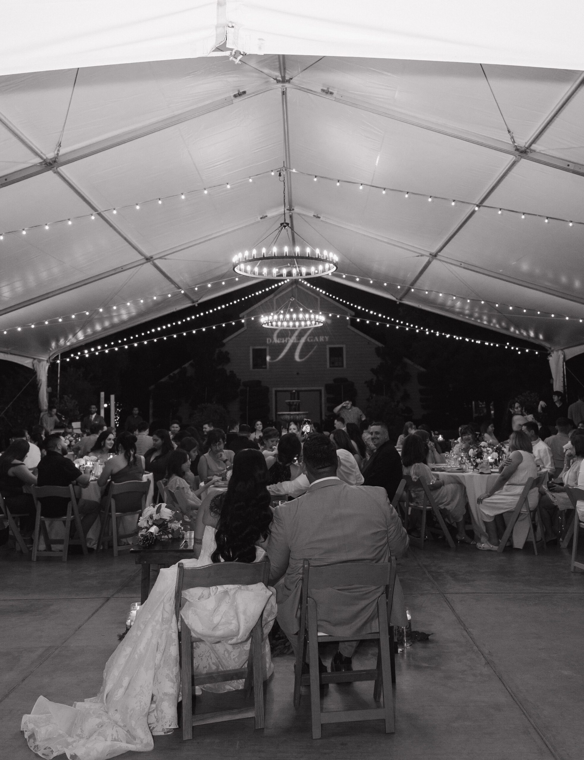 Black and white wedding photo of bride and groom sitting at sweetheart table looking at their wedding guests