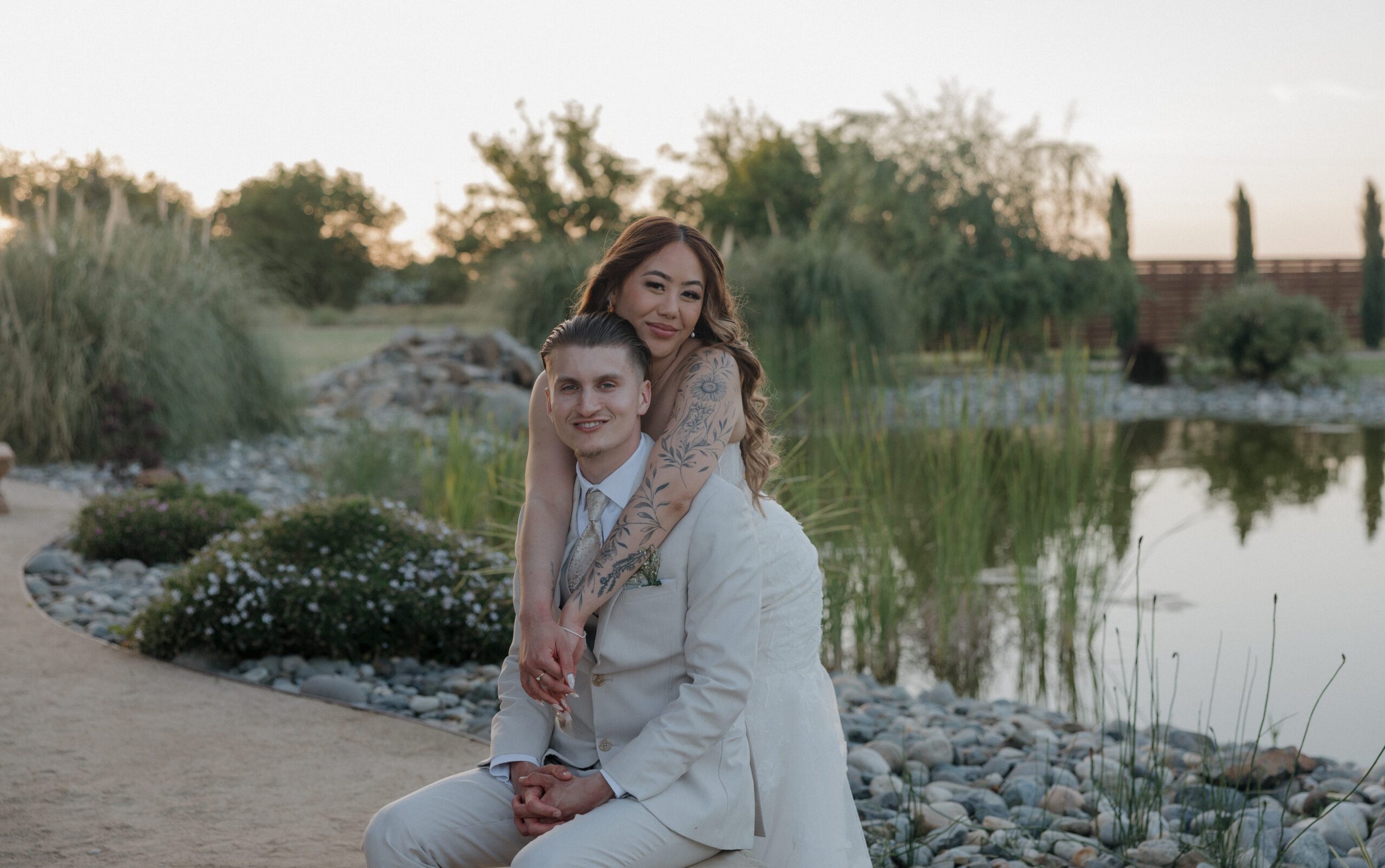 A bride hugging a groom from behind with a lake behind them during golden hour wedding photos