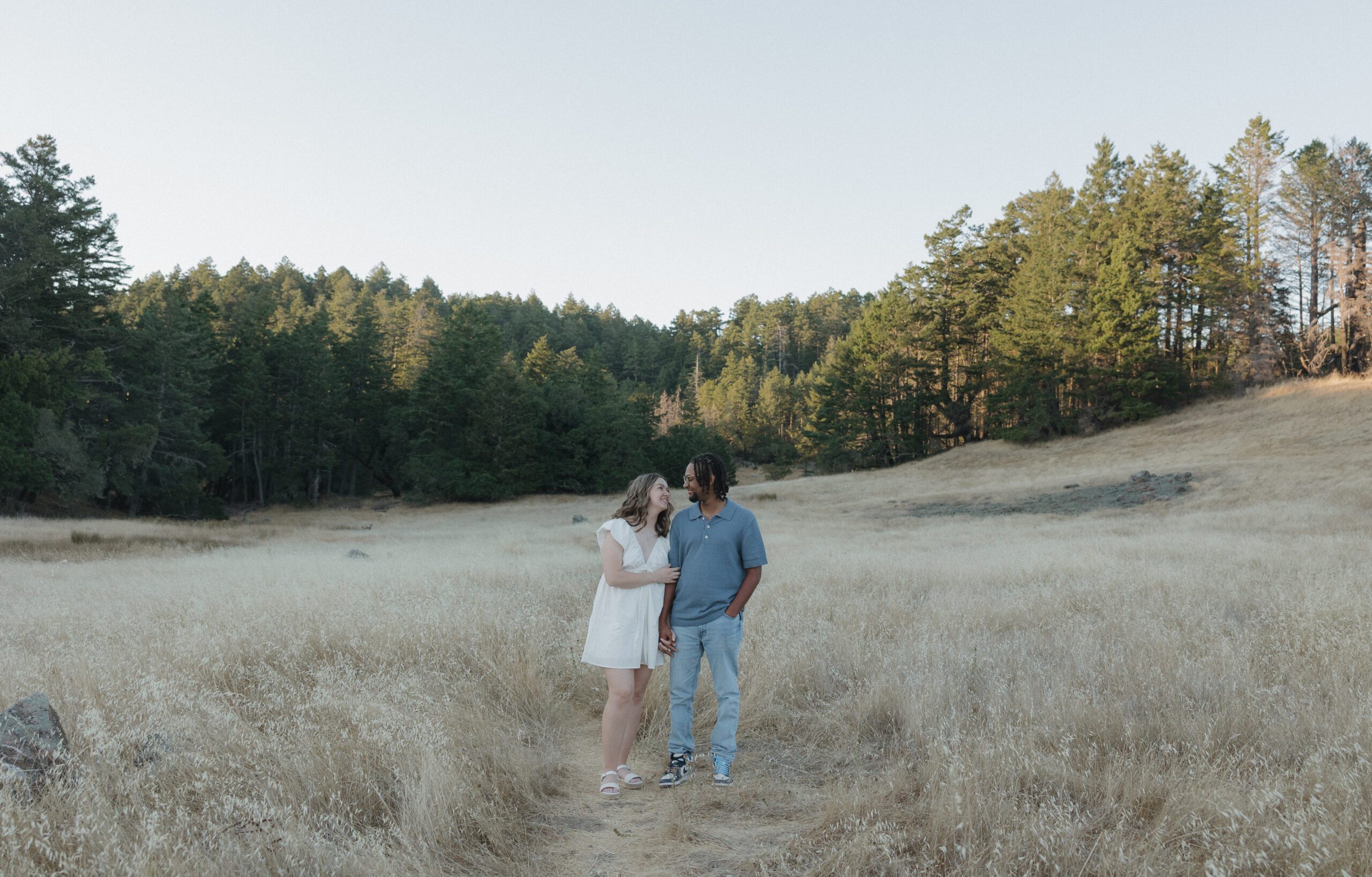 A couple smiling at each other while in a field and surrounded my trees at Mt. Tamalpais