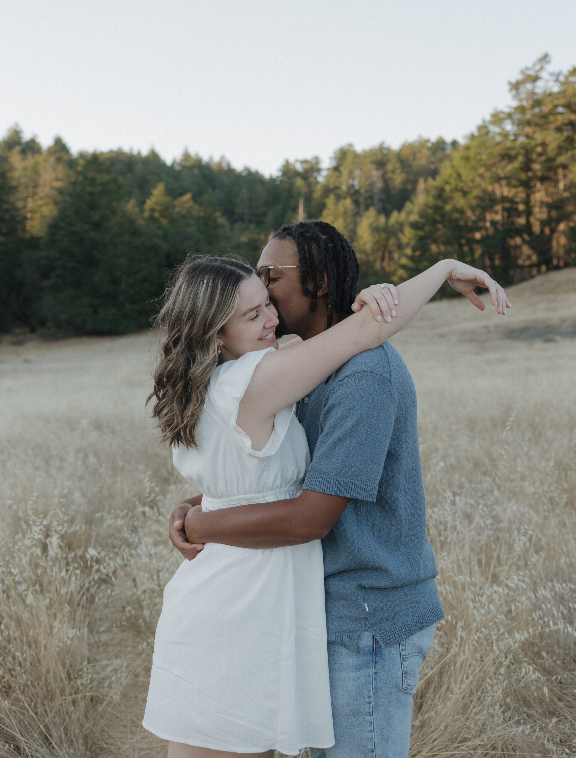 A man kissing his fiancee on the temple in a field at Mt. Tamalpais