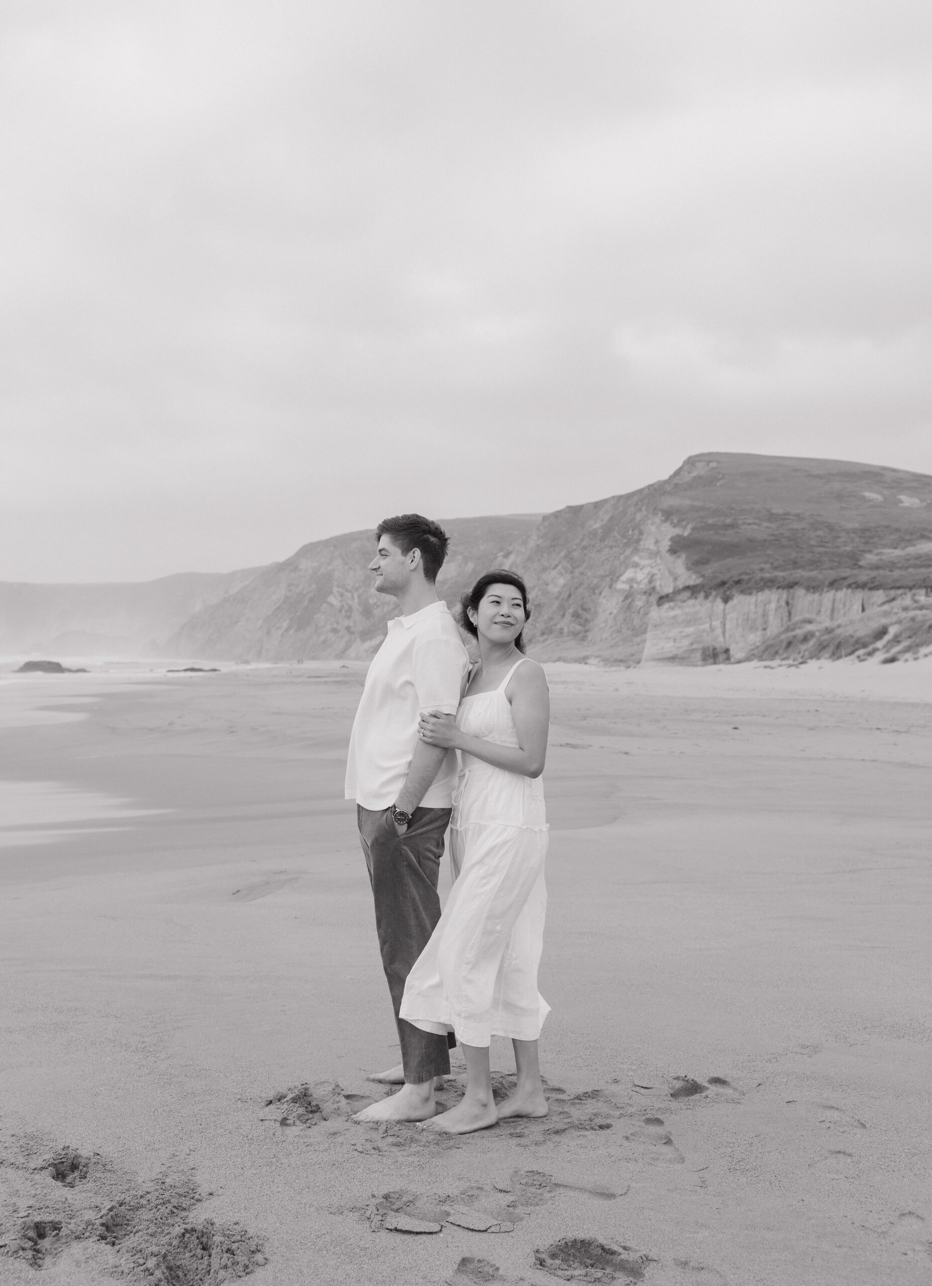 Black and white beach engagement photos of couple facing the ocean