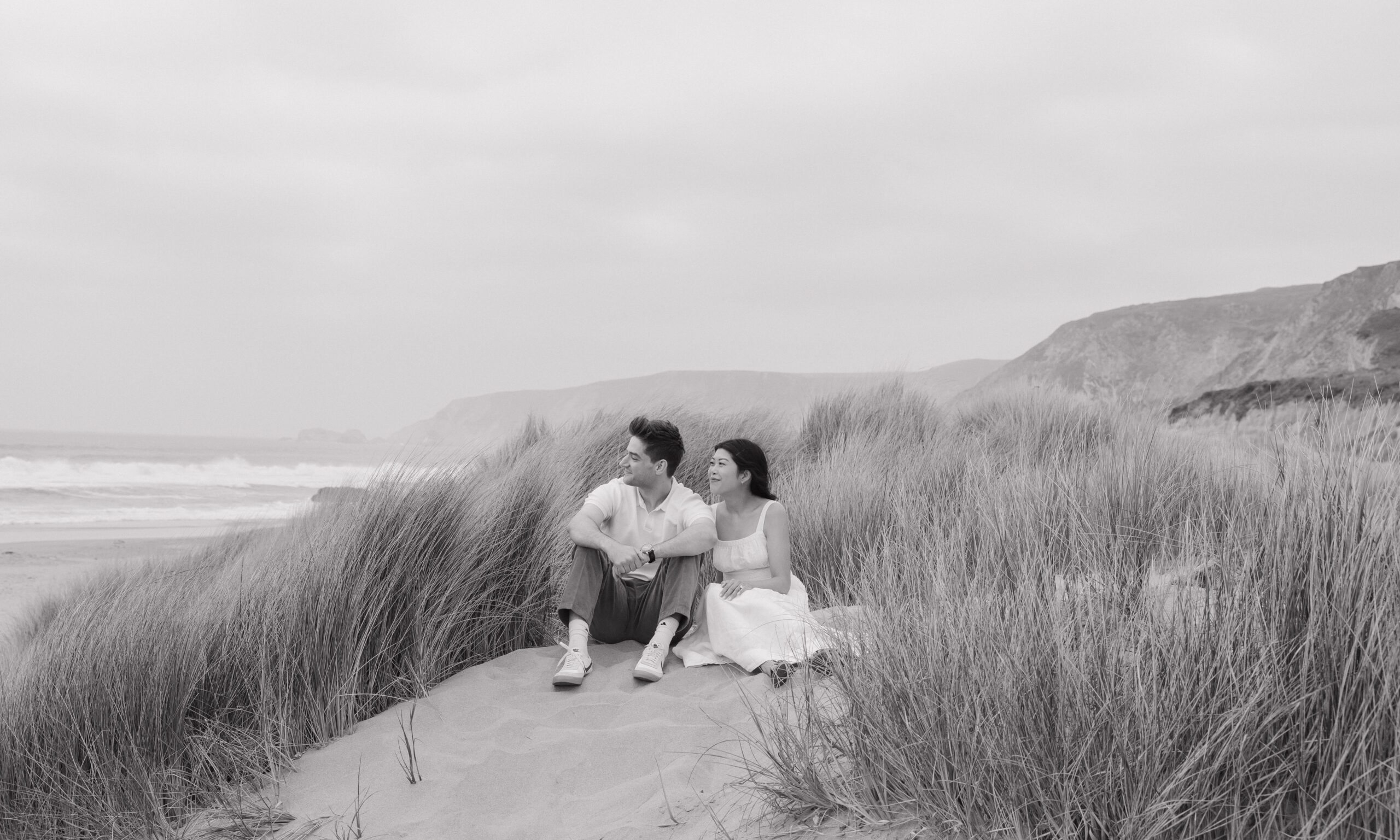 A black and white photo of a couple sitting in the sand during their engagement photos