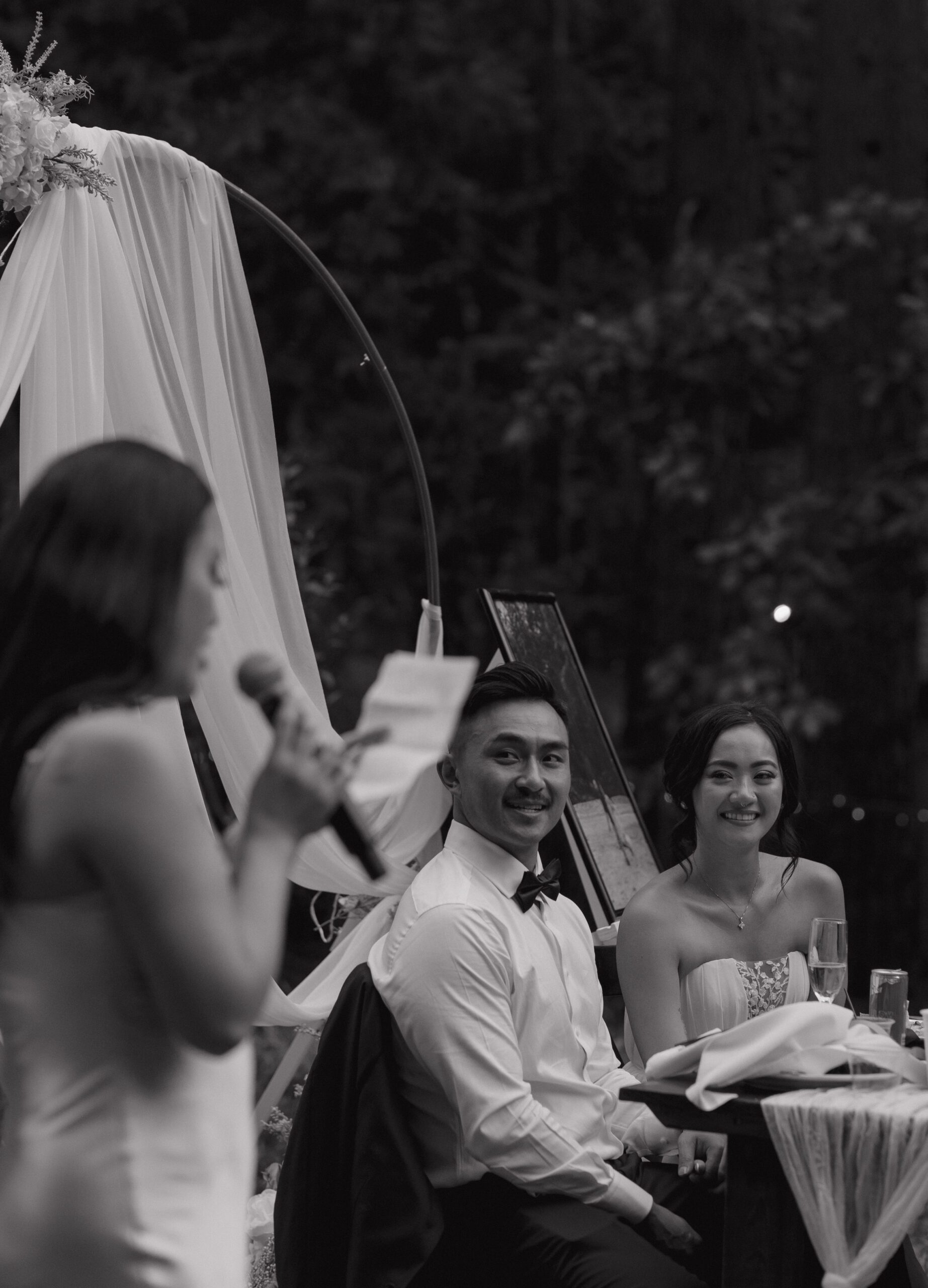 A candid wedding photo of a couple watching a bridesmaid give a toast