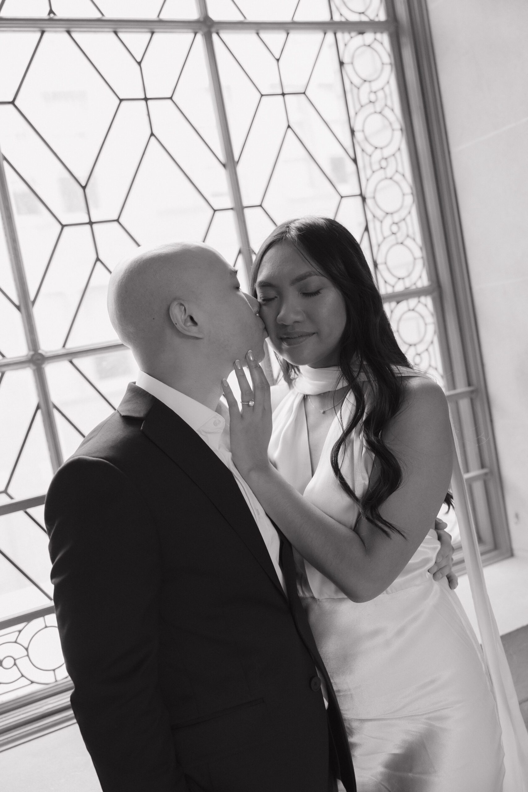 A black and white photo of a man kissing his fiancee on the cheek during their engagement photoshoot