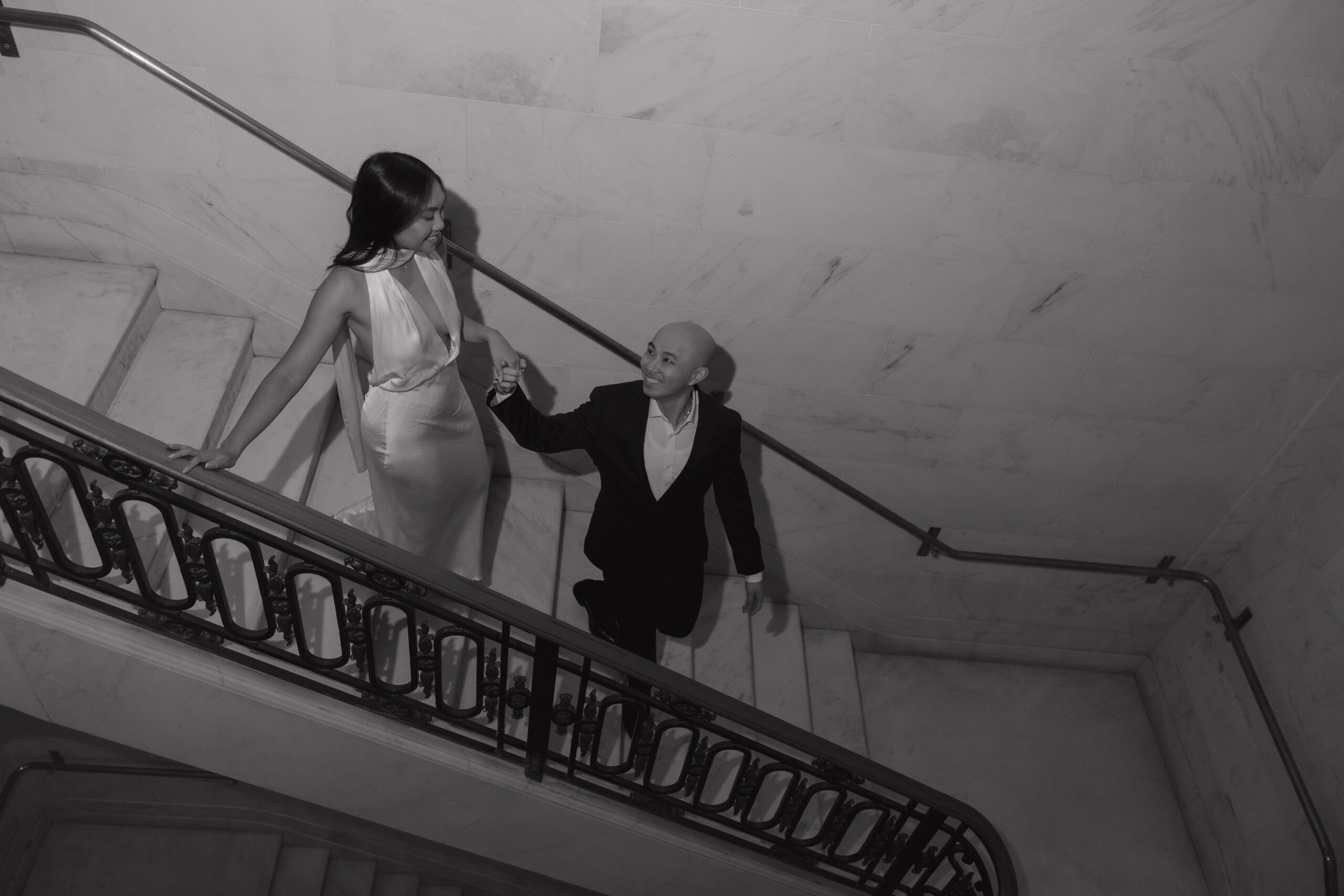 A black and white photo taken during a city hall engagement photoshoot of a man leading his fiancee down the stairs