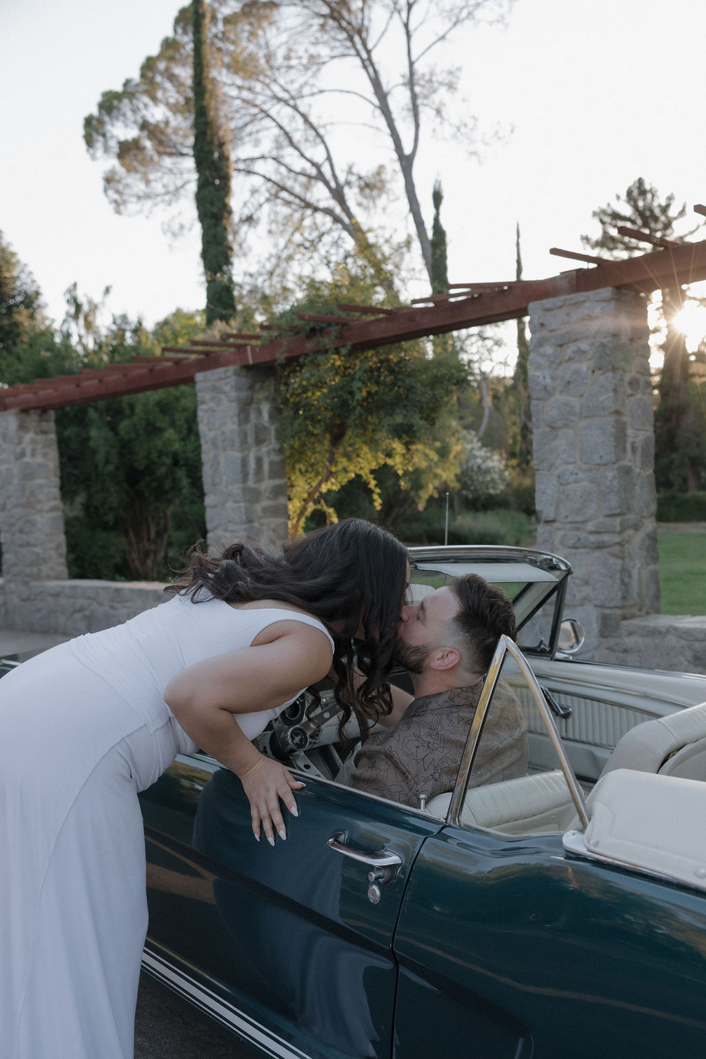 A woman leaning down to kiss her fiancé, who is in a classic car.