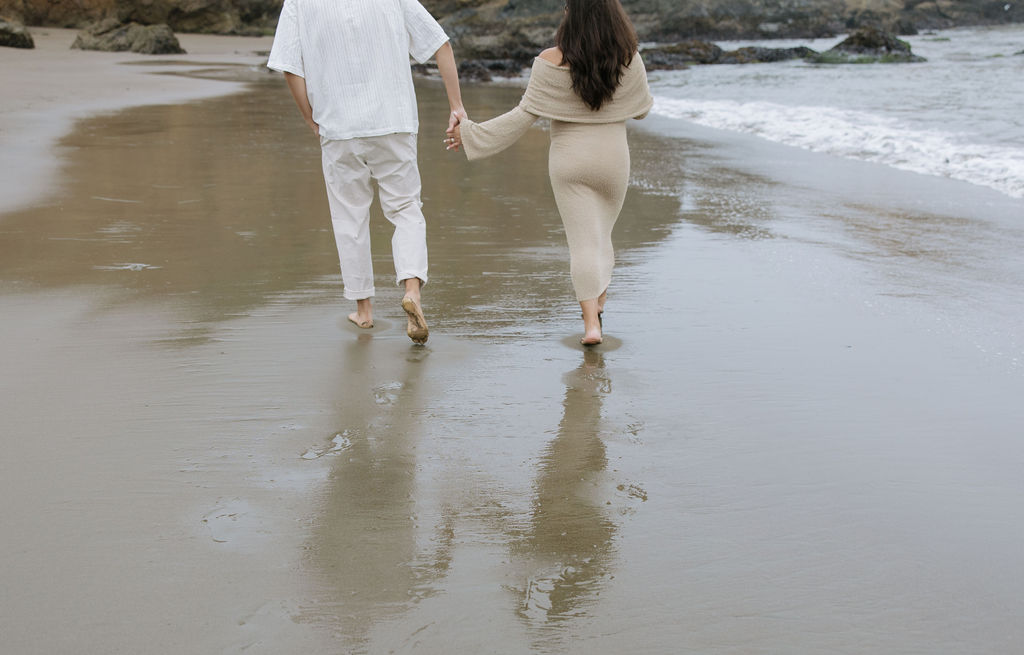 A woman holding hands with her husband walking along the ocean for maternity photos