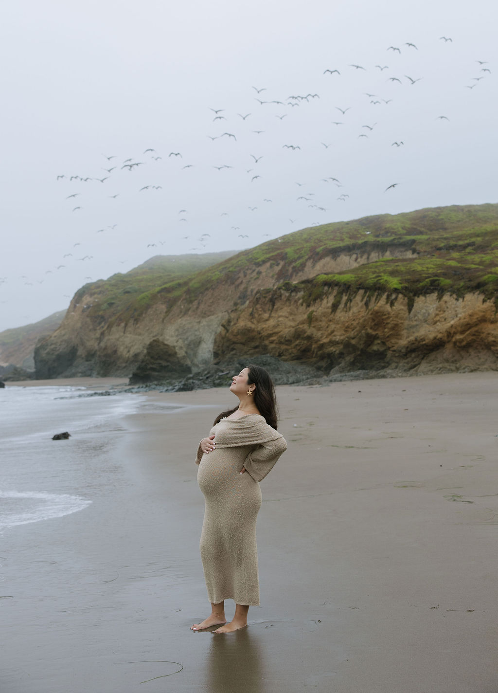 A woman posing for beach maternity photos with birds flying behind her