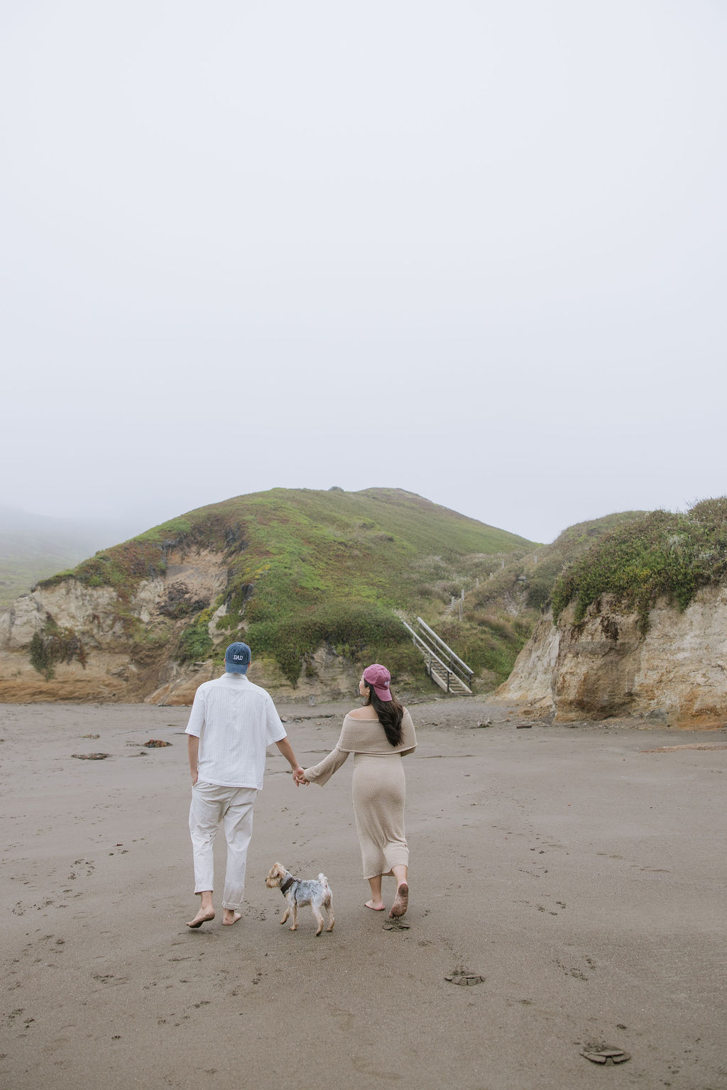 A couple holding hands walking on the beach wearing mom and dad hats for a maternity photoshoot