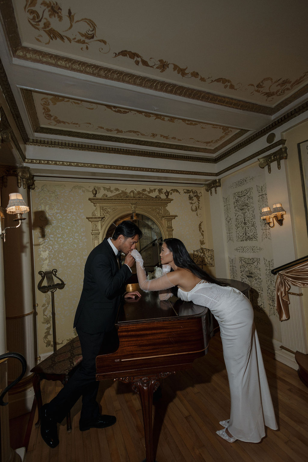A bride leaning over a piano while her groom kisses her hand inside of grand island mansion