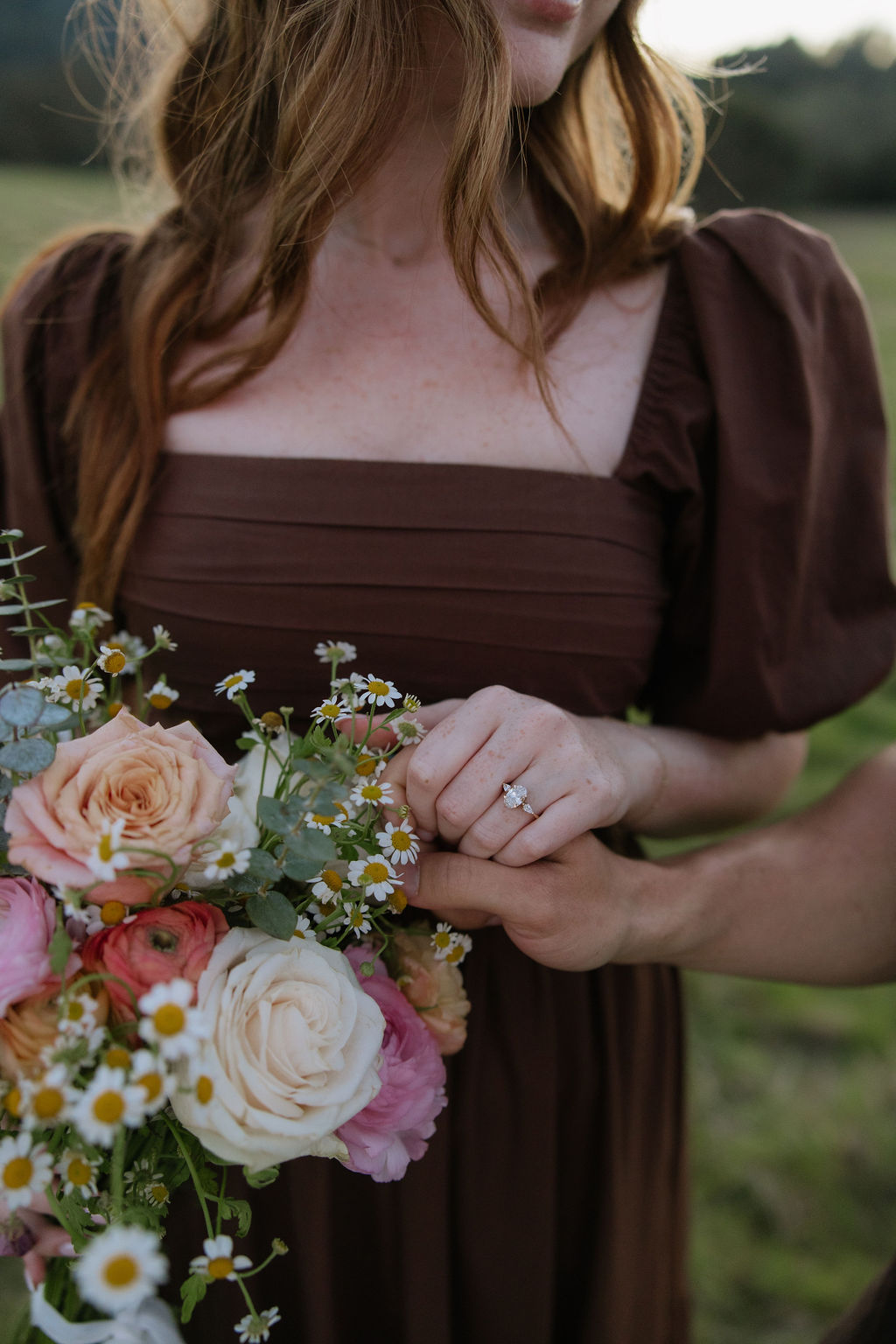 A woman in a brown dress showing off her bouquet and engagement ring