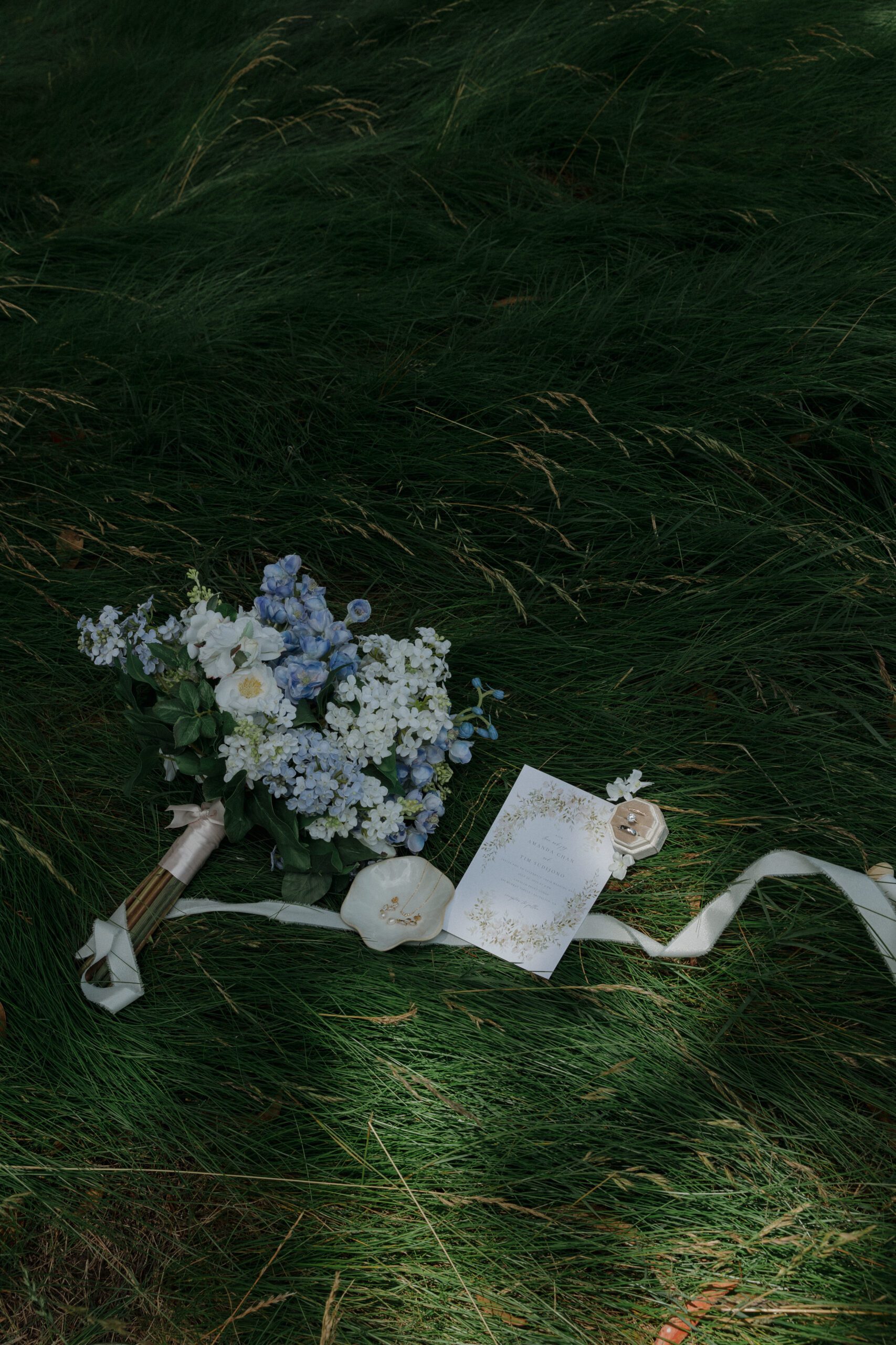 A wedding flat lay with a blue bridal bouquet, rings, and invitations