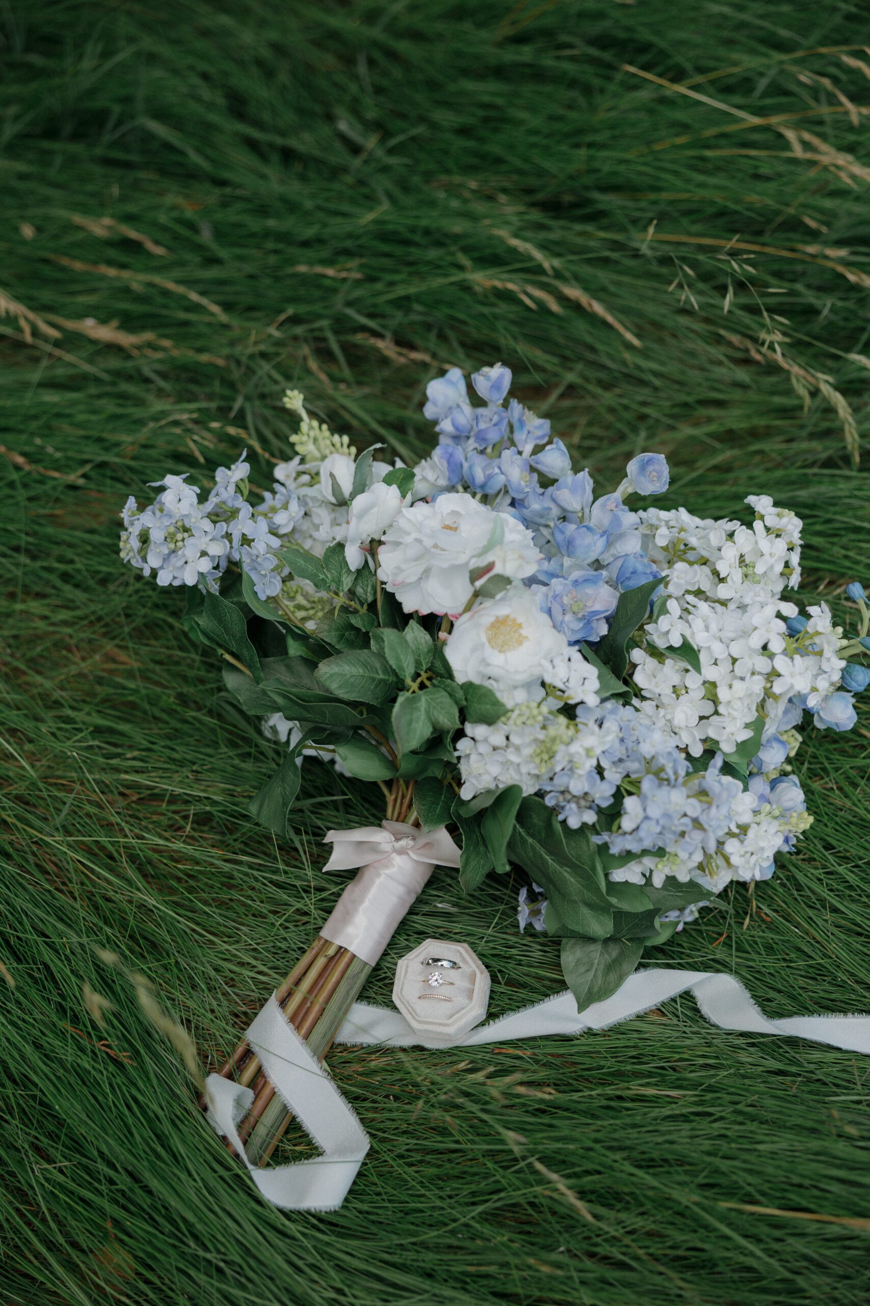 A wedding flat lay with a blue bridal bouquet, rings, and invitations